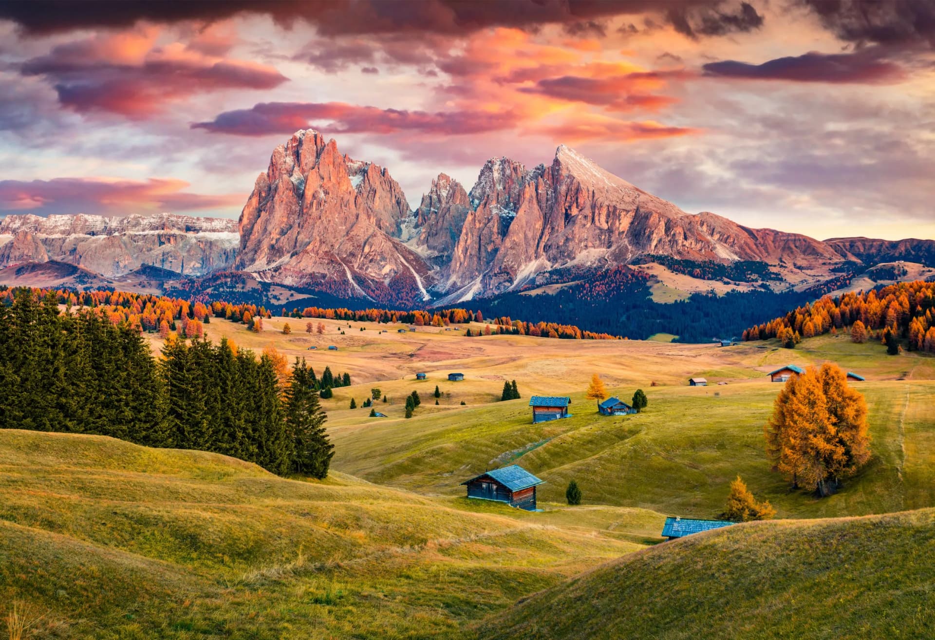 Alpe di Siusi mountain range with autumn foliage, rolling hills, and small wooden huts at sunset.