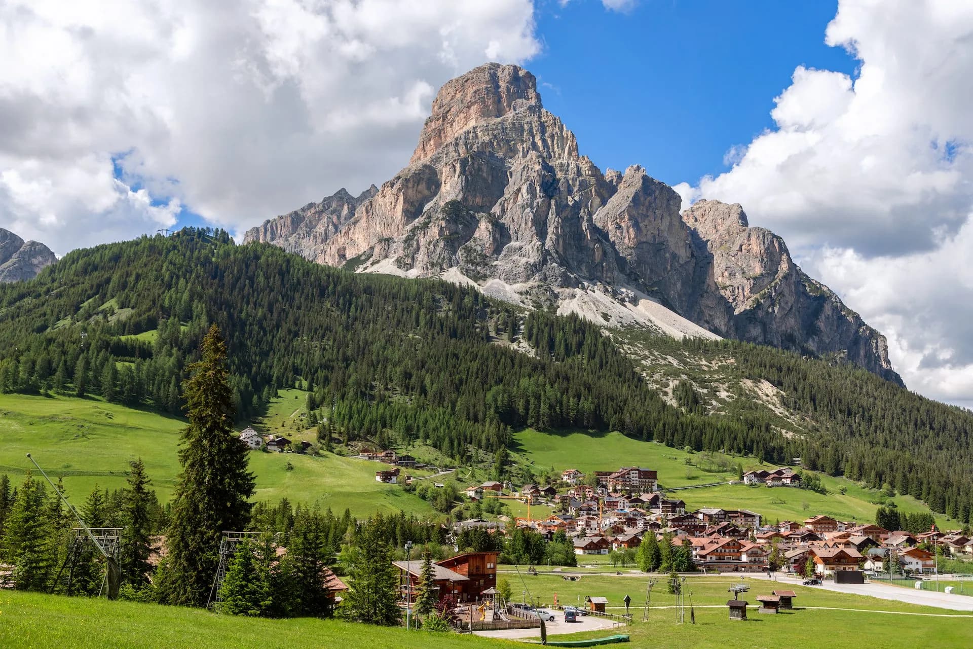 Alpine village nestled in green valley below massive rocky mountain under blue sky with clouds.