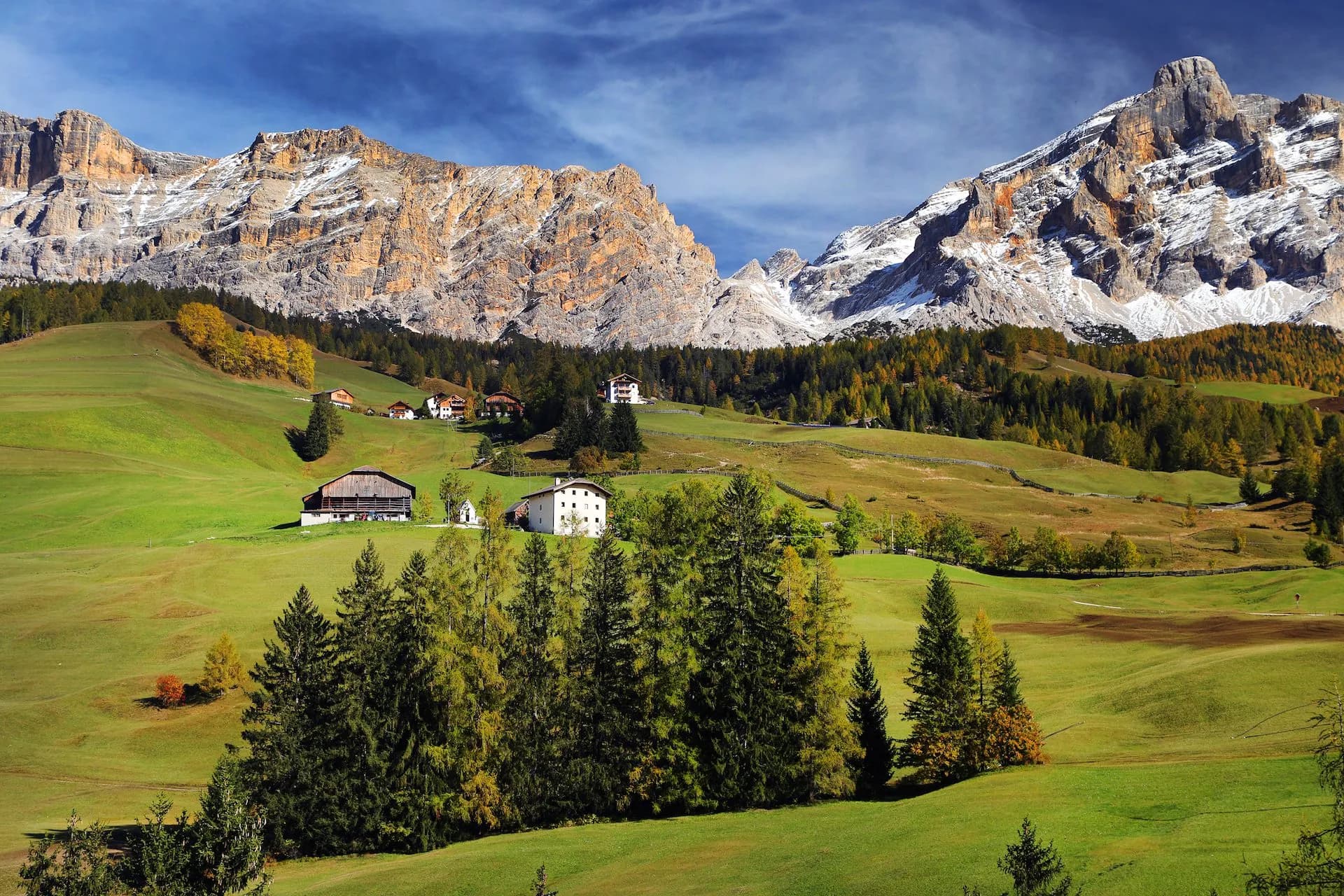Alpine village houses nestled in green meadows beneath snow-dusted Dolomites mountains in autumn.