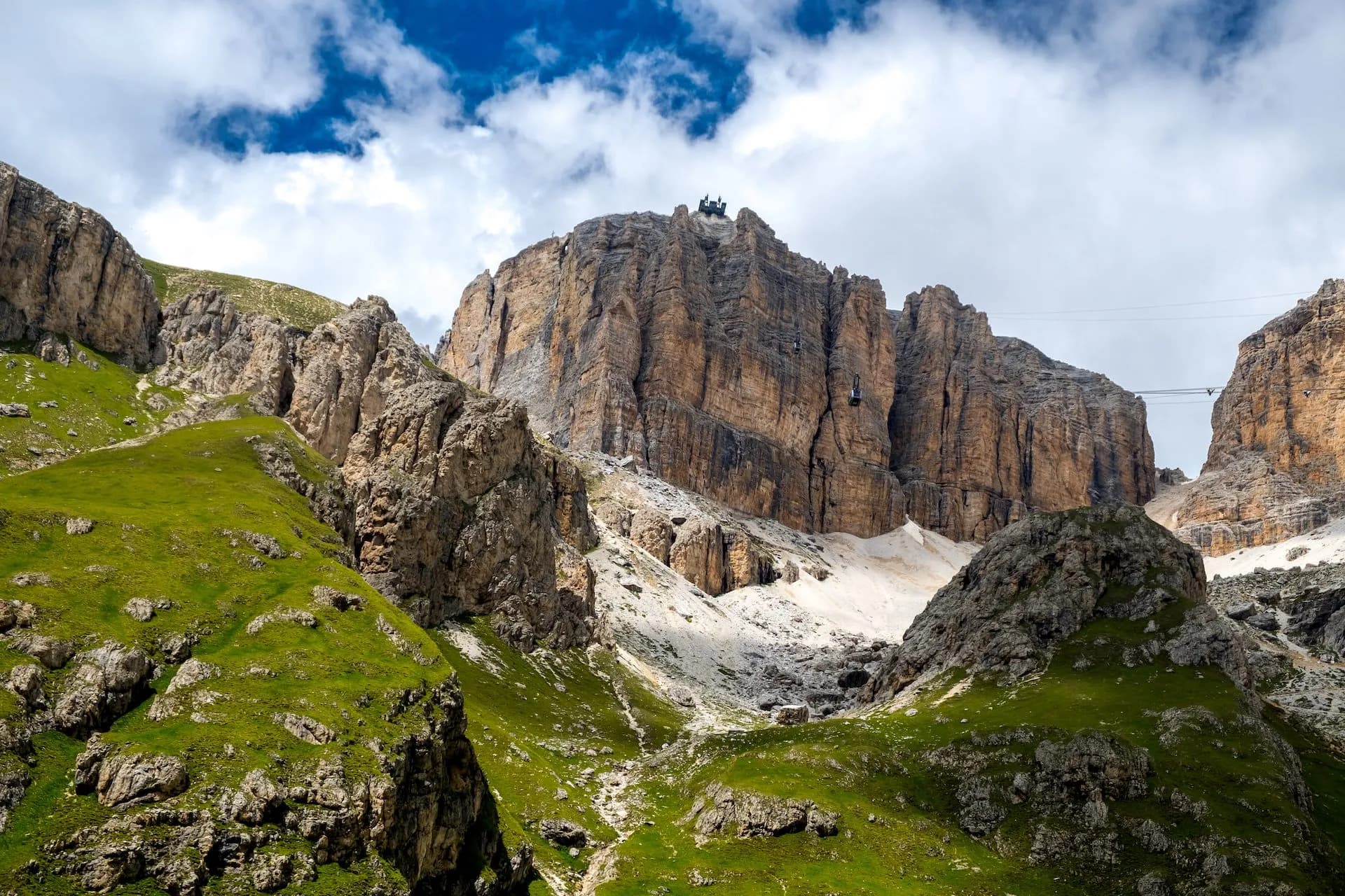 Cable car ascending towering rocky mountains above green alpine slopes under a cloudy sky.