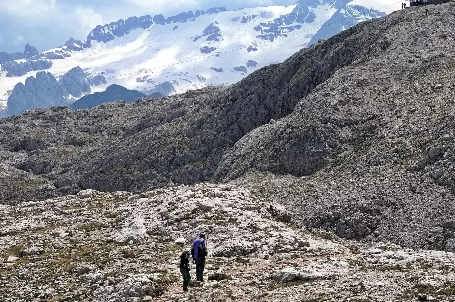 Hikers on rocky terrain below snow-capped mountains near Rifugio Franz Kostner