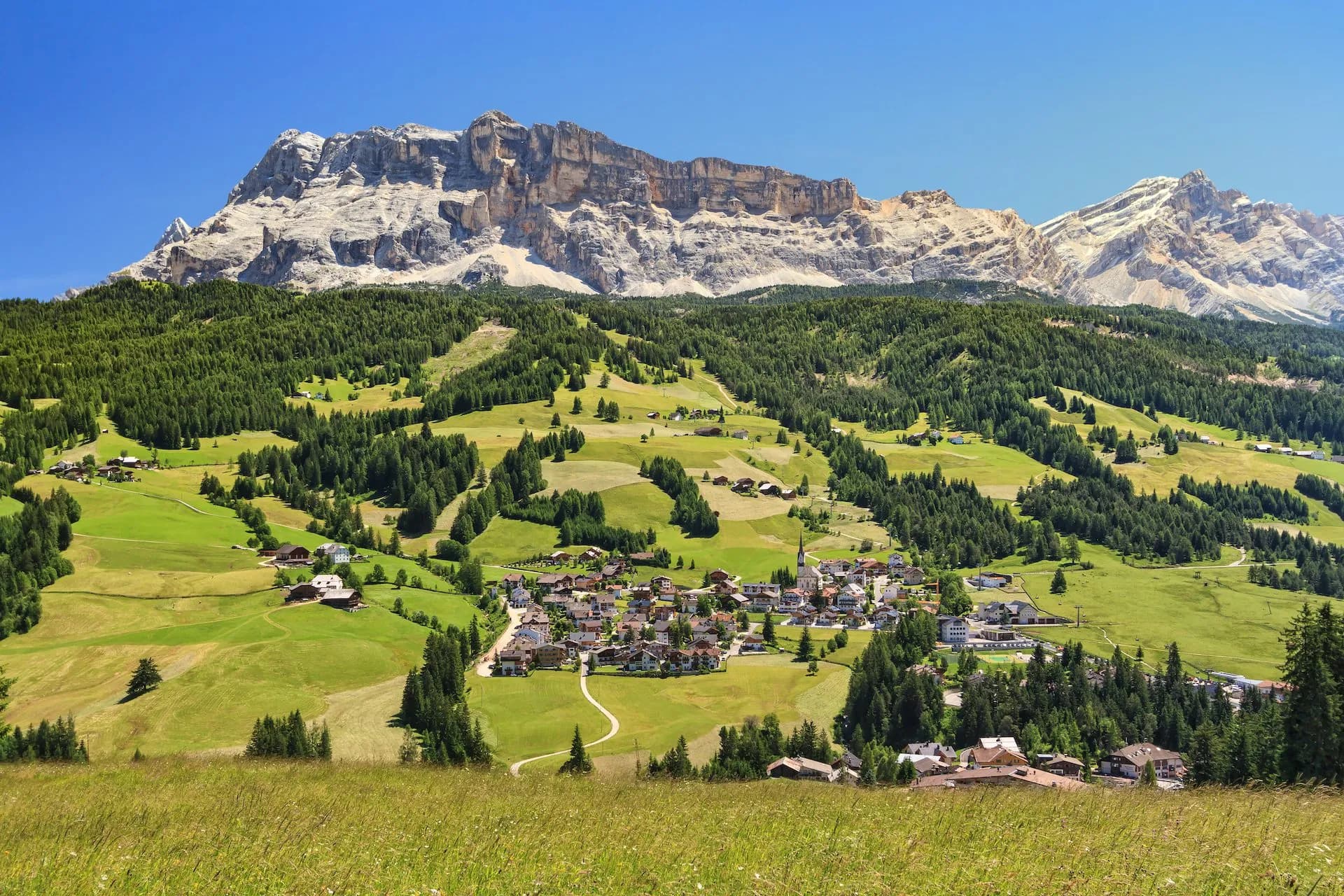 Alpine village nestled in green valley below towering, rocky mountains under clear blue sky in Badia.