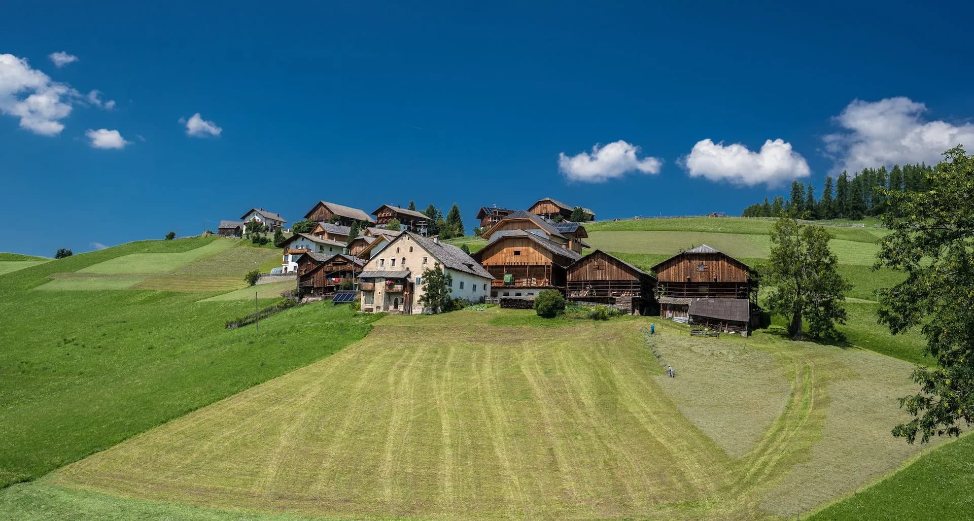 Alpine village houses on green hillside under bright blue sky with scattered white clouds, La Val in Badia.