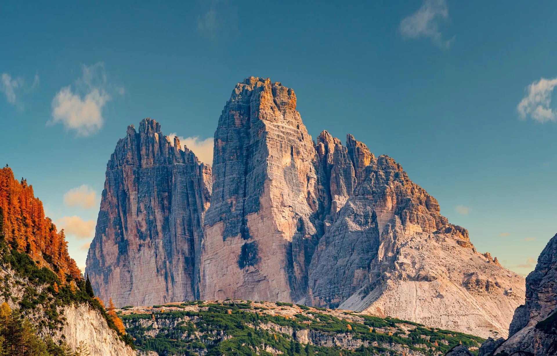 Tre Cime di Lavaredo mountains illuminated by sunlight with autumn foliage under a blue sky.
