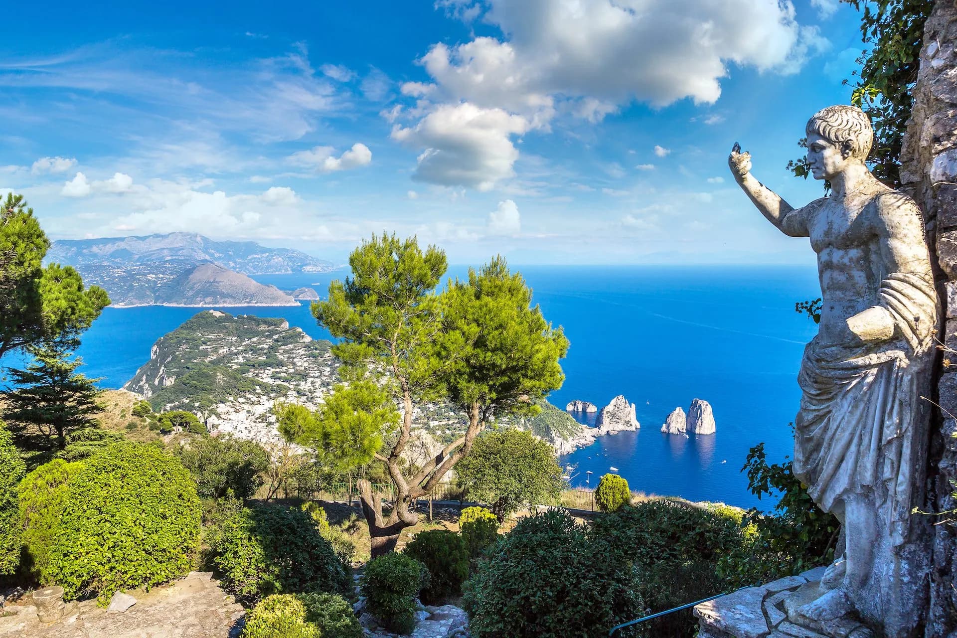 Statue overlooking Capri Faraglioni rocks, lush gardens, and bright blue Mediterranean Sea.