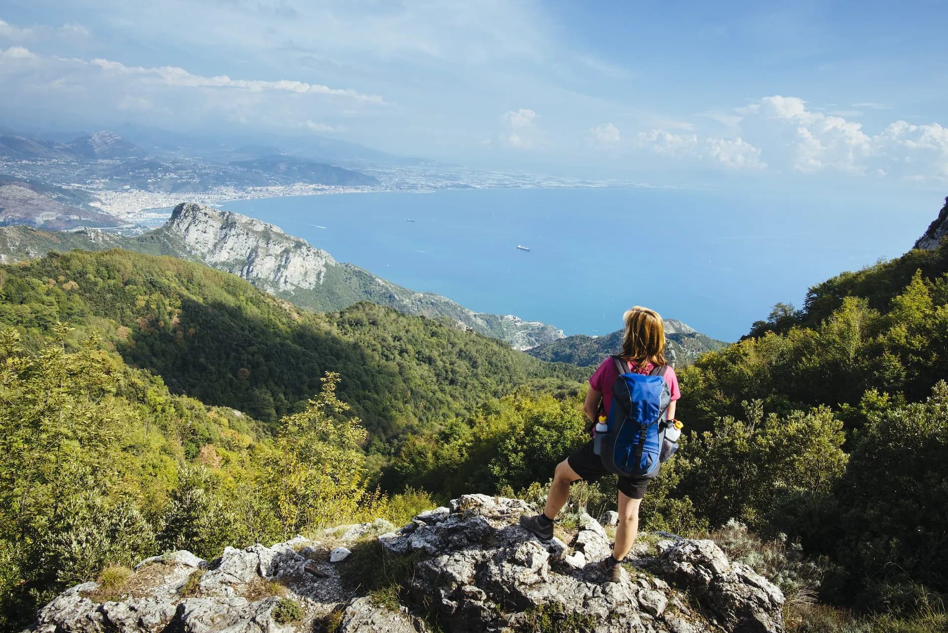 Hiker with backpack overlooking lush green mountains and coastal city on bright blue sea.