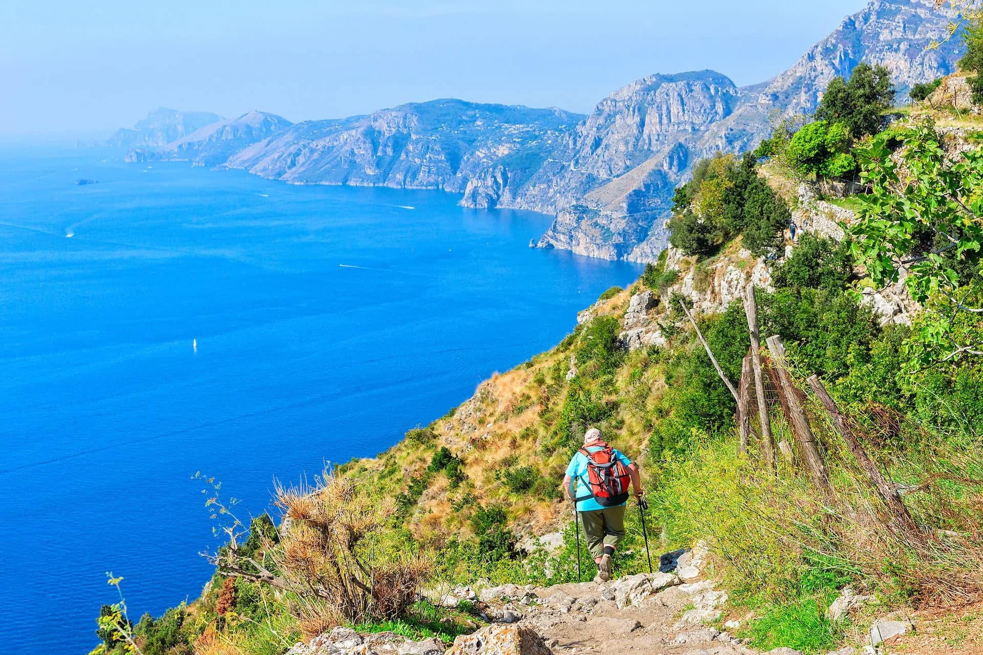 Hiker with poles on coastal path overlooking bright blue sea and rugged mountains.