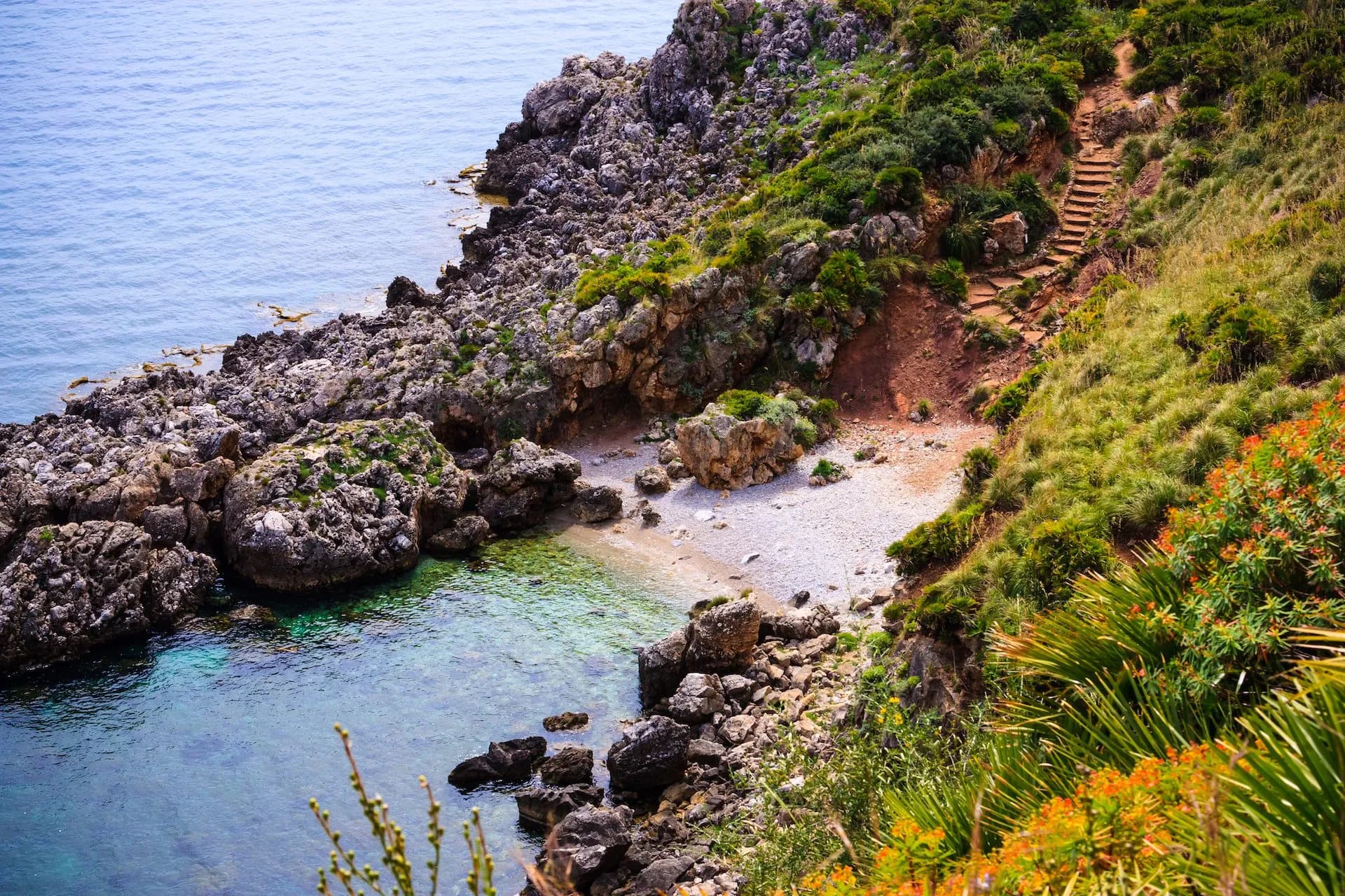 Rocky coastline with small cove, clear blue water, and hiking steps in Zingaro Nature Reserve, Sicily.