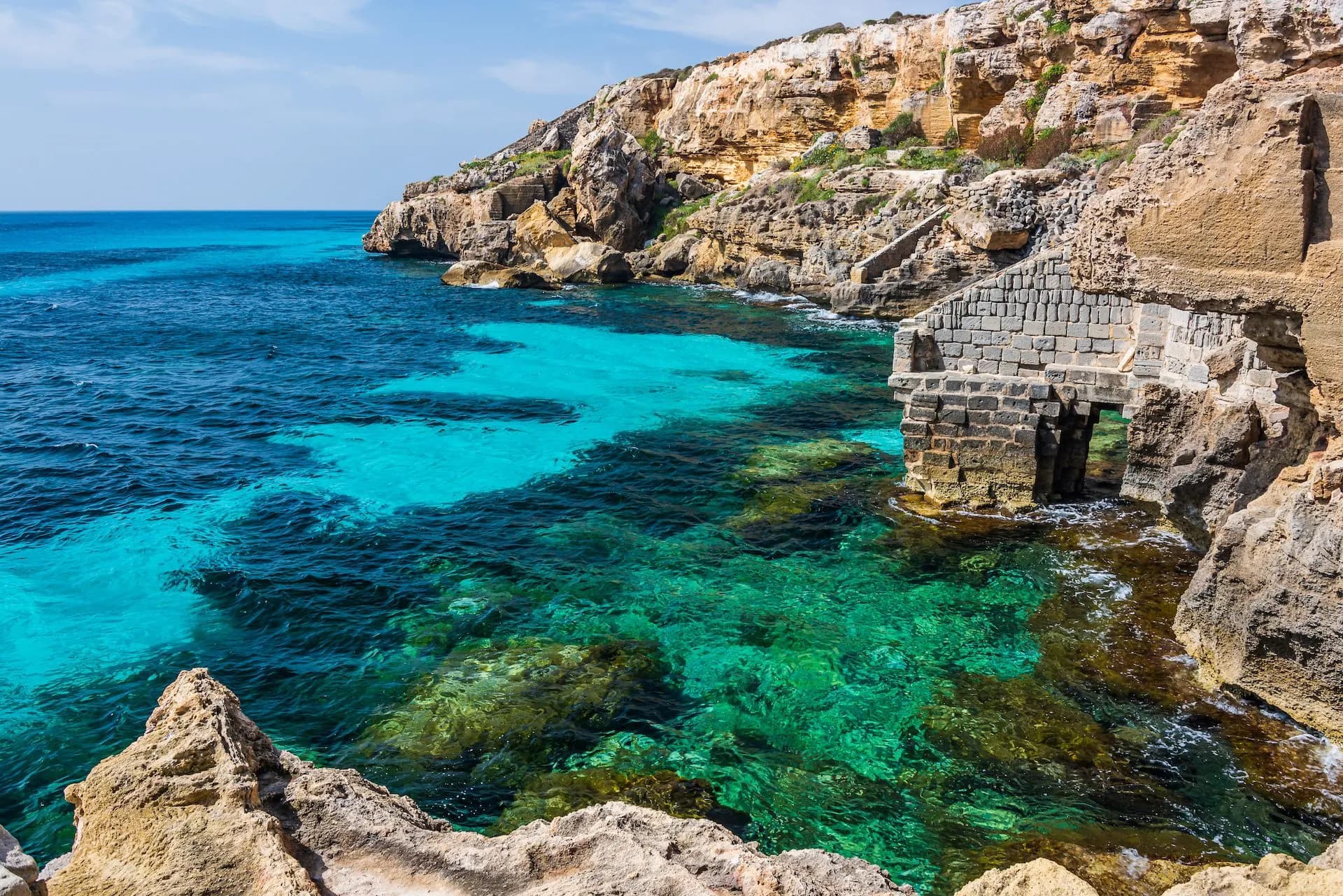 Turquoise water meets rocky cliffs and a stone structure on the Favignana coastline.
