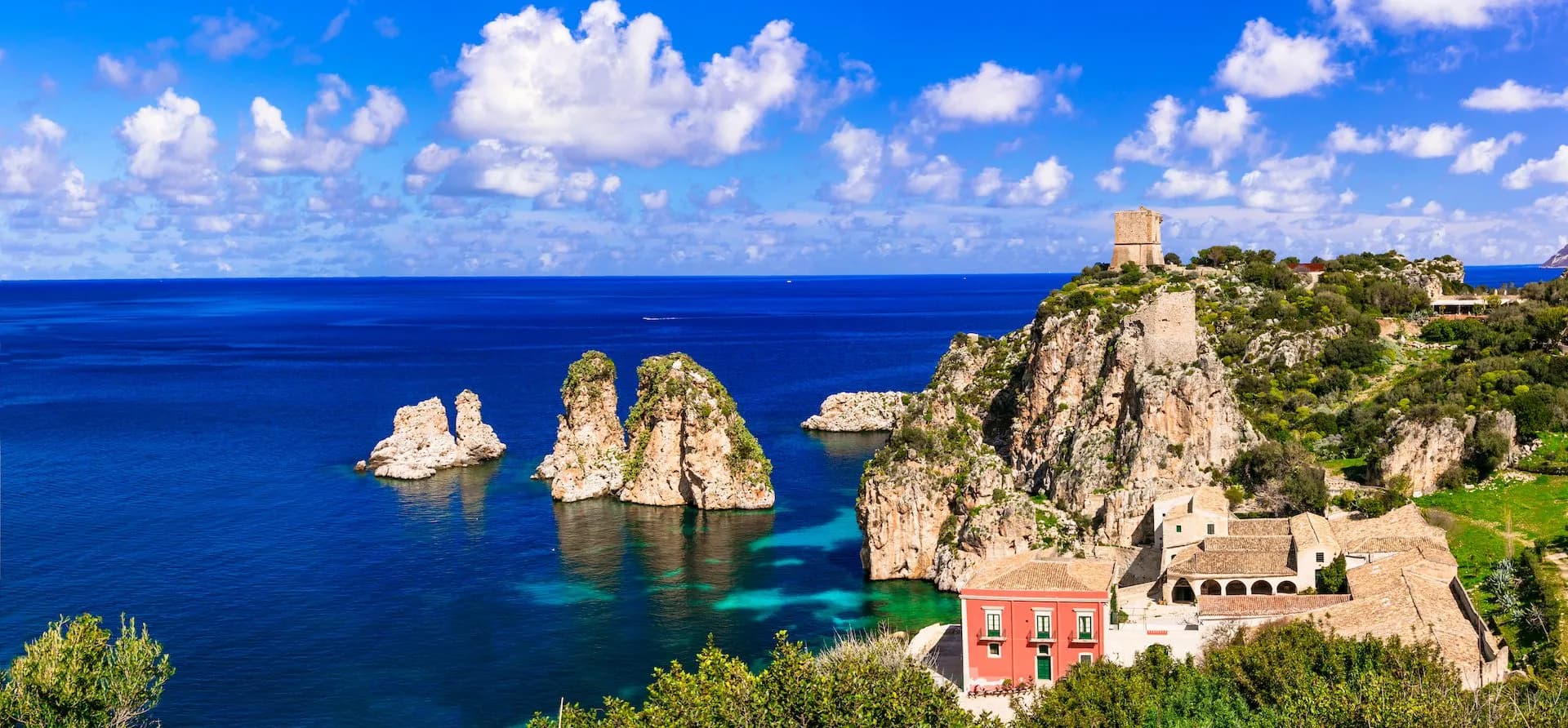 Coastal view of Scopello with sea stacks, historic tower, and buildings above turquoise water.