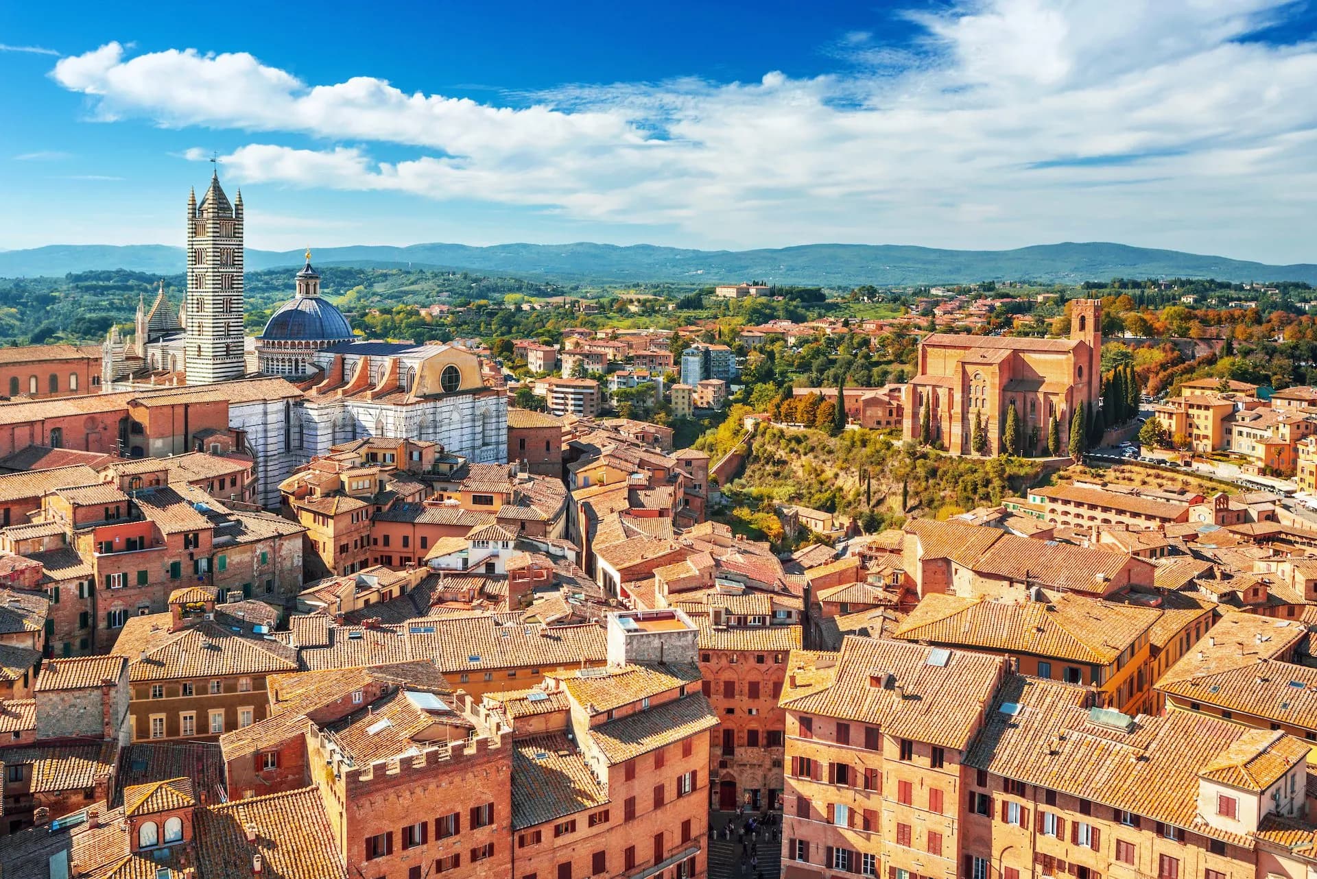 Rooftops of medieval Siena with the striped Duomo tower and surrounding Tuscan hills under a blue sky.