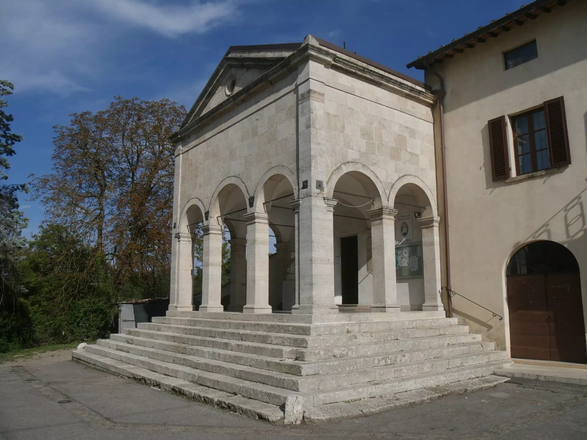 Stone church portico with arches and steps, next to a building under a blue sky in Gracciano d'Elsa.