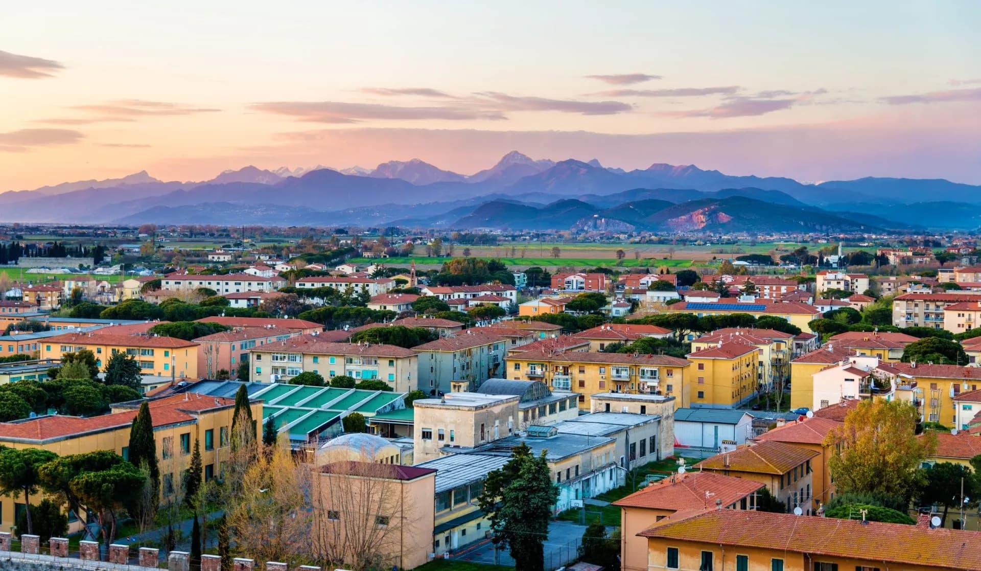 City rooftops with terracotta tiles and Apuan Alps mountains in the distance at sunset.