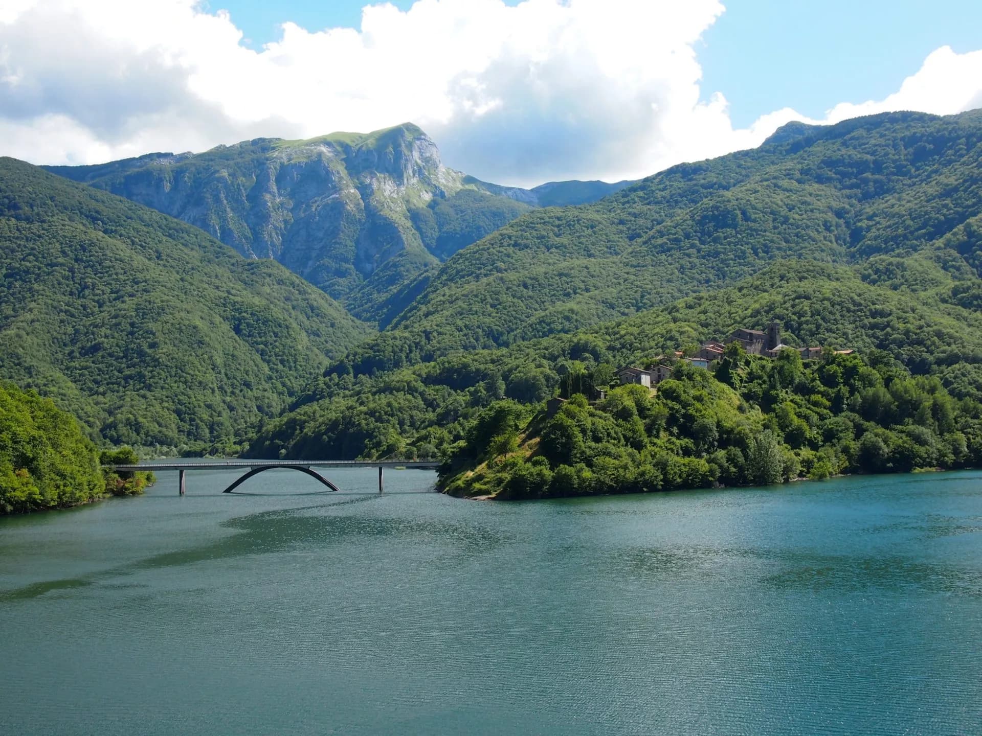 Arch bridge over Lago di Vagli reservoir surrounded by lush green mountains under a cloudy sky.