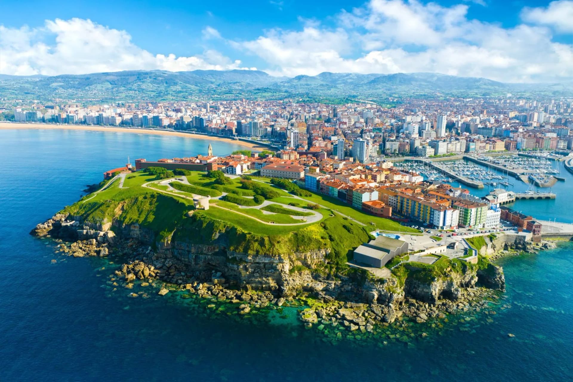 Aerial view of Gijón, Asturias, showing a city, green cliffside park, harbor, and mountains under a blue sky.