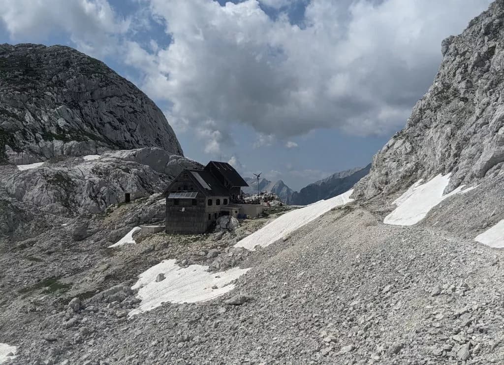 Mountain lodge nestled in rocky terrain with patches of snow under a cloudy sky, Dolič Mountain.