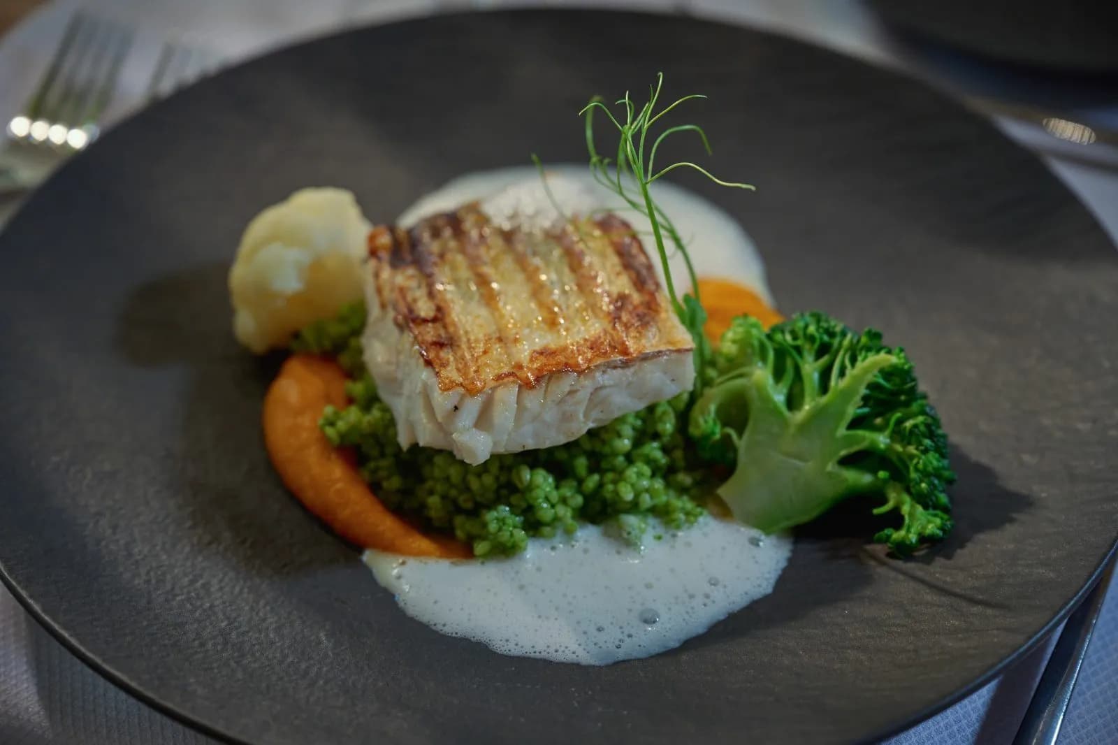 Grilled fish fillet with green couscous, broccoli, cauliflower, and foam sauce on a dark plate.