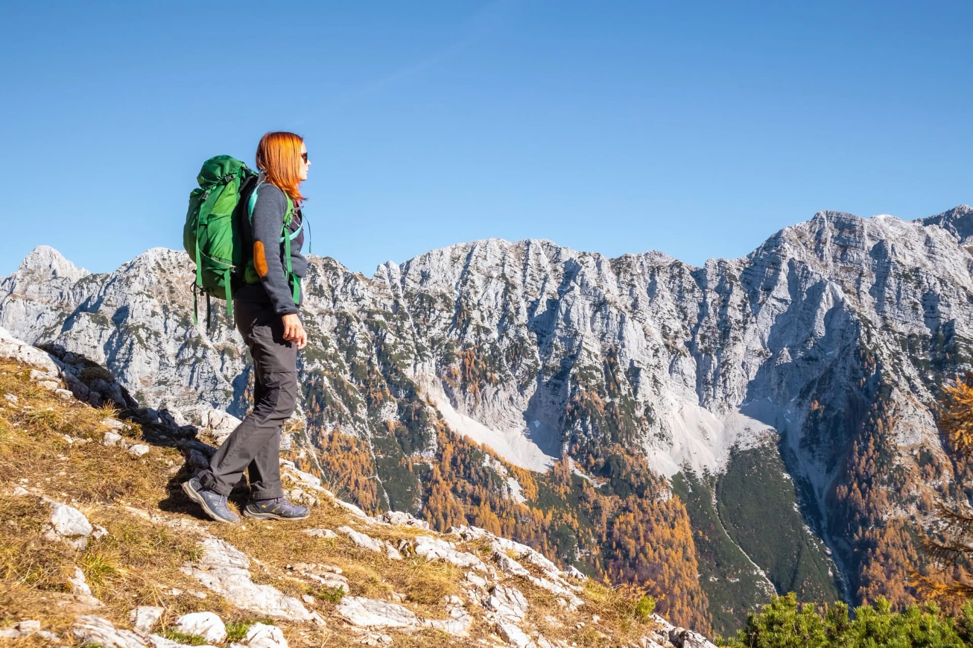 Hiker with green backpack overlooking Julian Alps mountains with autumn foliage under clear blue sky.
