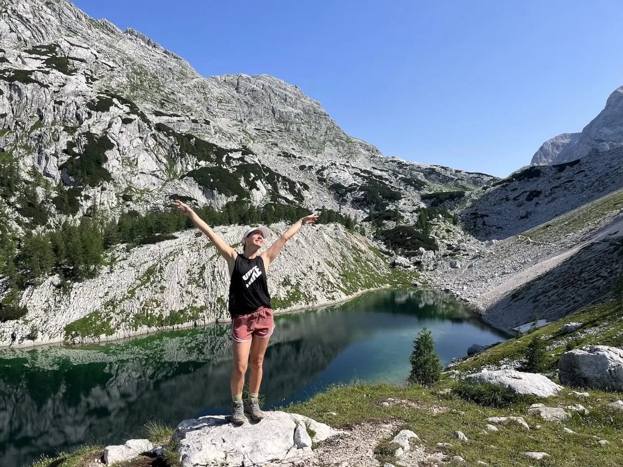 Hiker with arms raised by a green alpine lake surrounded by rocky mountains under a clear blue sky.