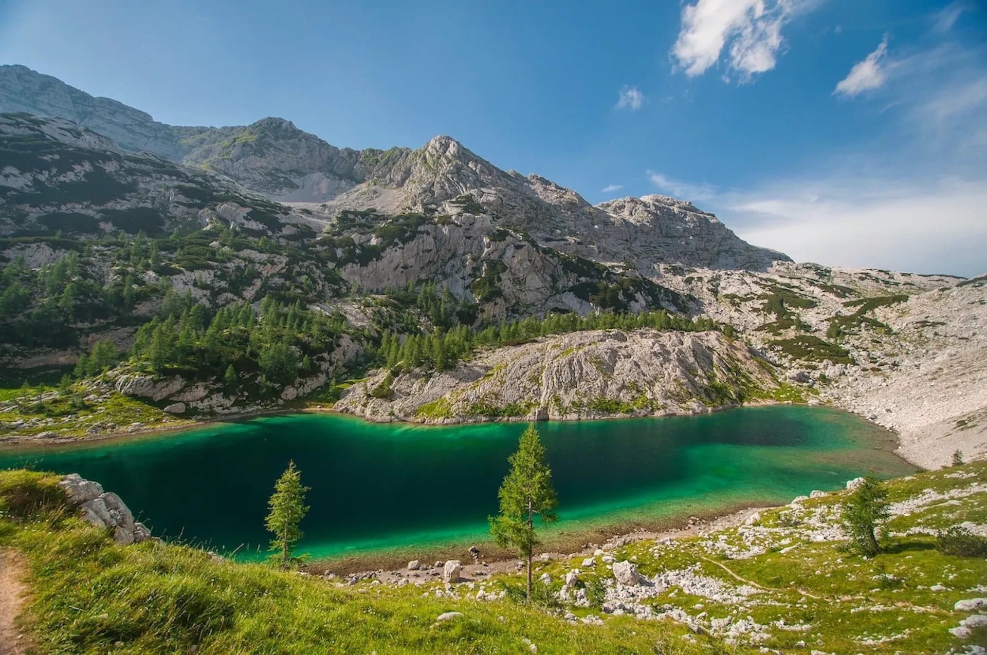 Alpine lake with emerald water surrounded by rocky mountains and green vegetation under a blue sky.