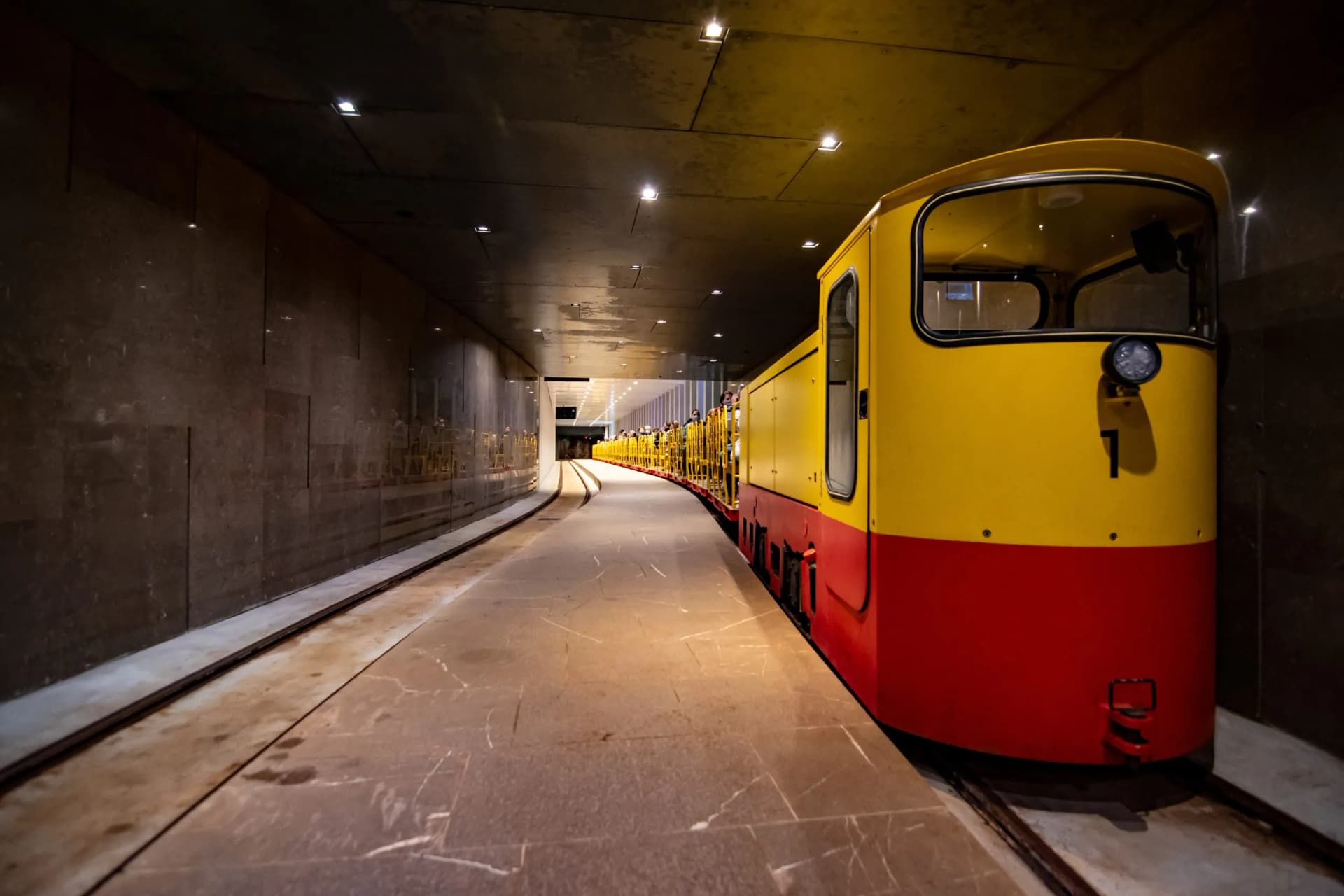 Yellow and red tourist train stopped in a dimly lit underground station with stone walls.