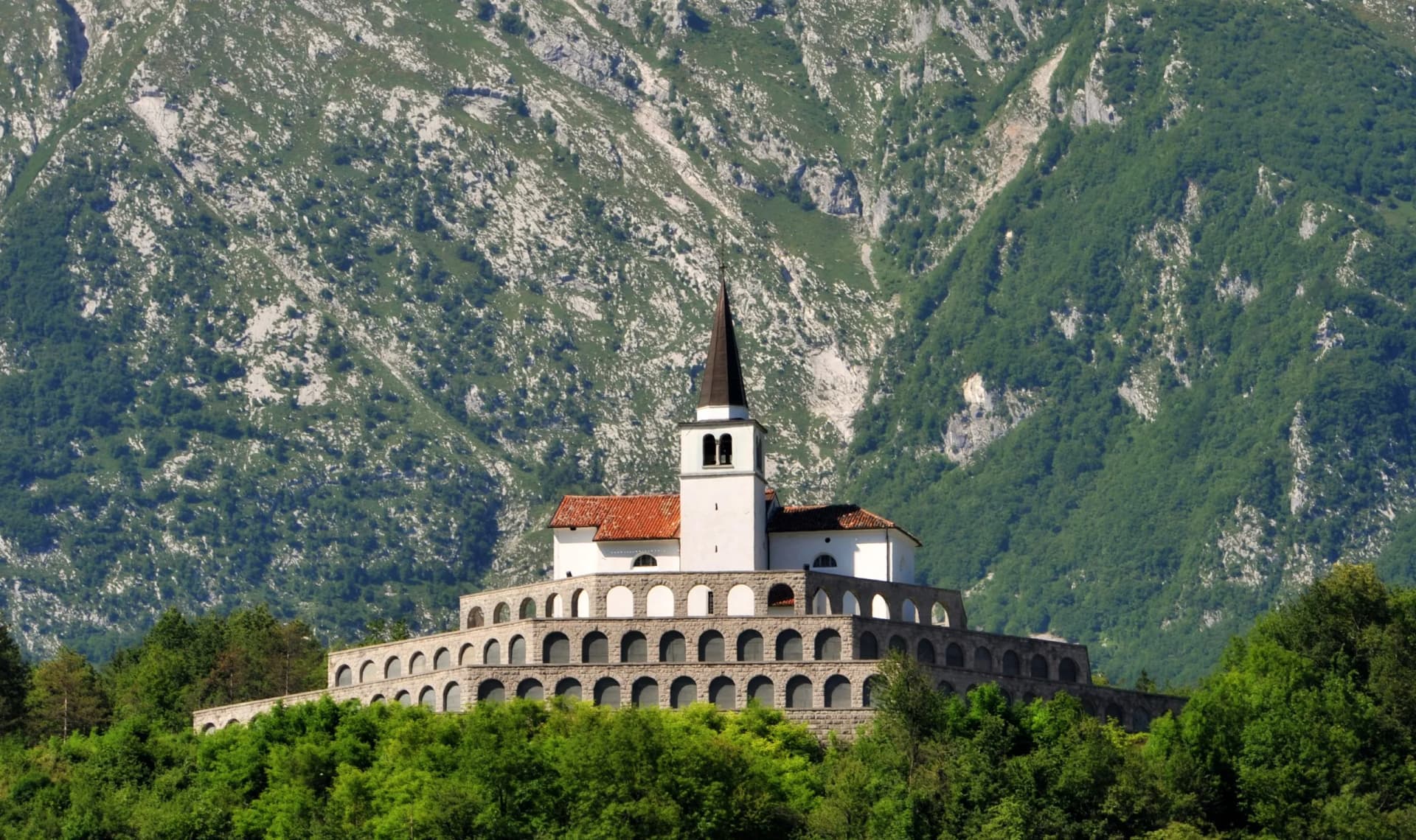 White church with bell tower on arched stone terrace against steep, green, rocky mountainside.