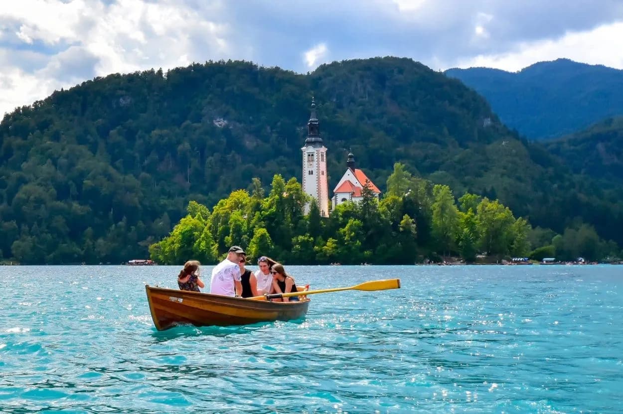 Rowing boat on turquoise Lake Bled toward island church with forested mountains backdrop