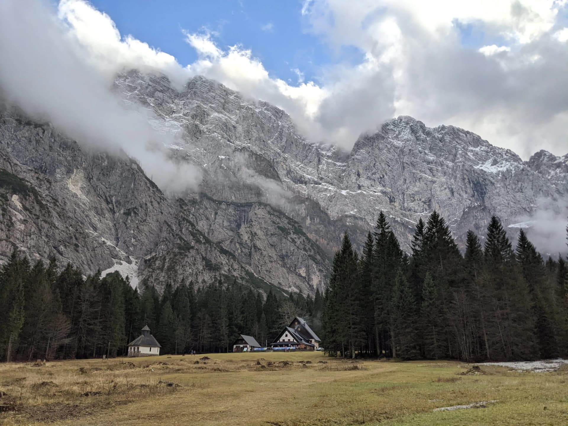 Mountain lodge and small chapel in a meadow below towering, cloud-shrouded rocky peaks.