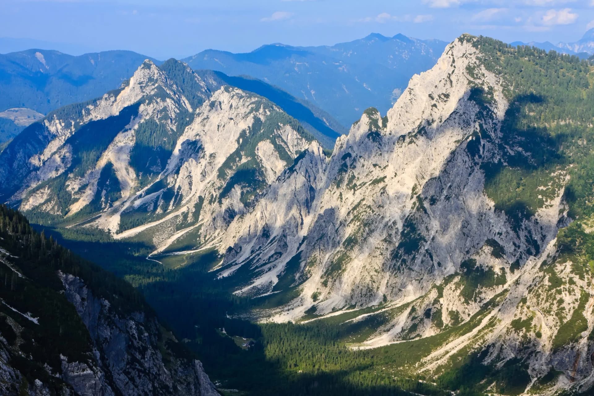 Rugged, sunlit mountainsides with steep, pale rock faces and dark green forests under a blue sky.