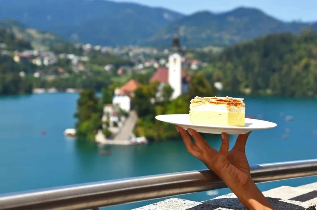Cream cake slice held over Lake Bled with island church and mountains in background