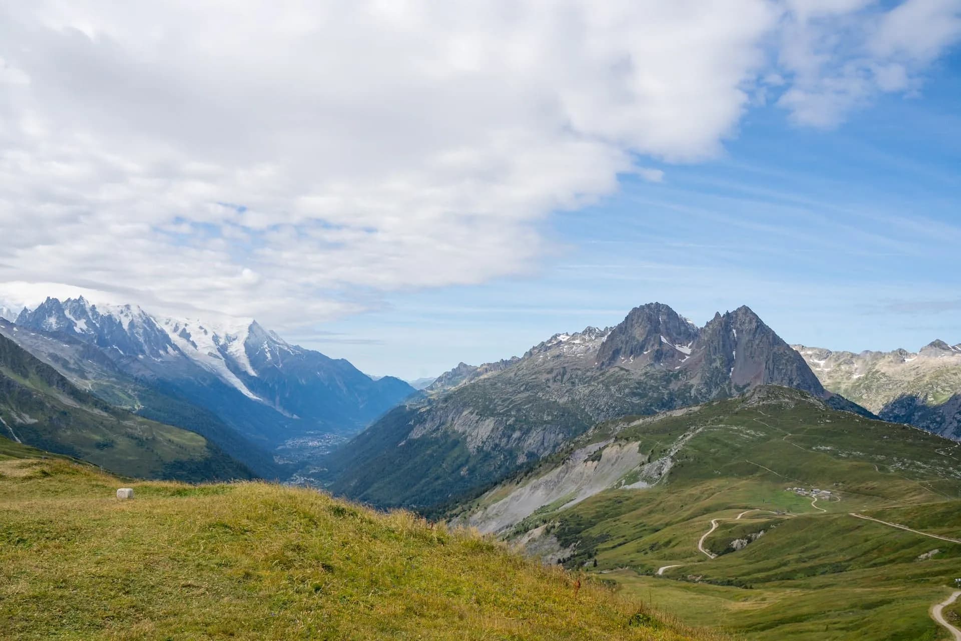 Mountain pass view with grassy foreground, snow-capped peaks, and valley town below.