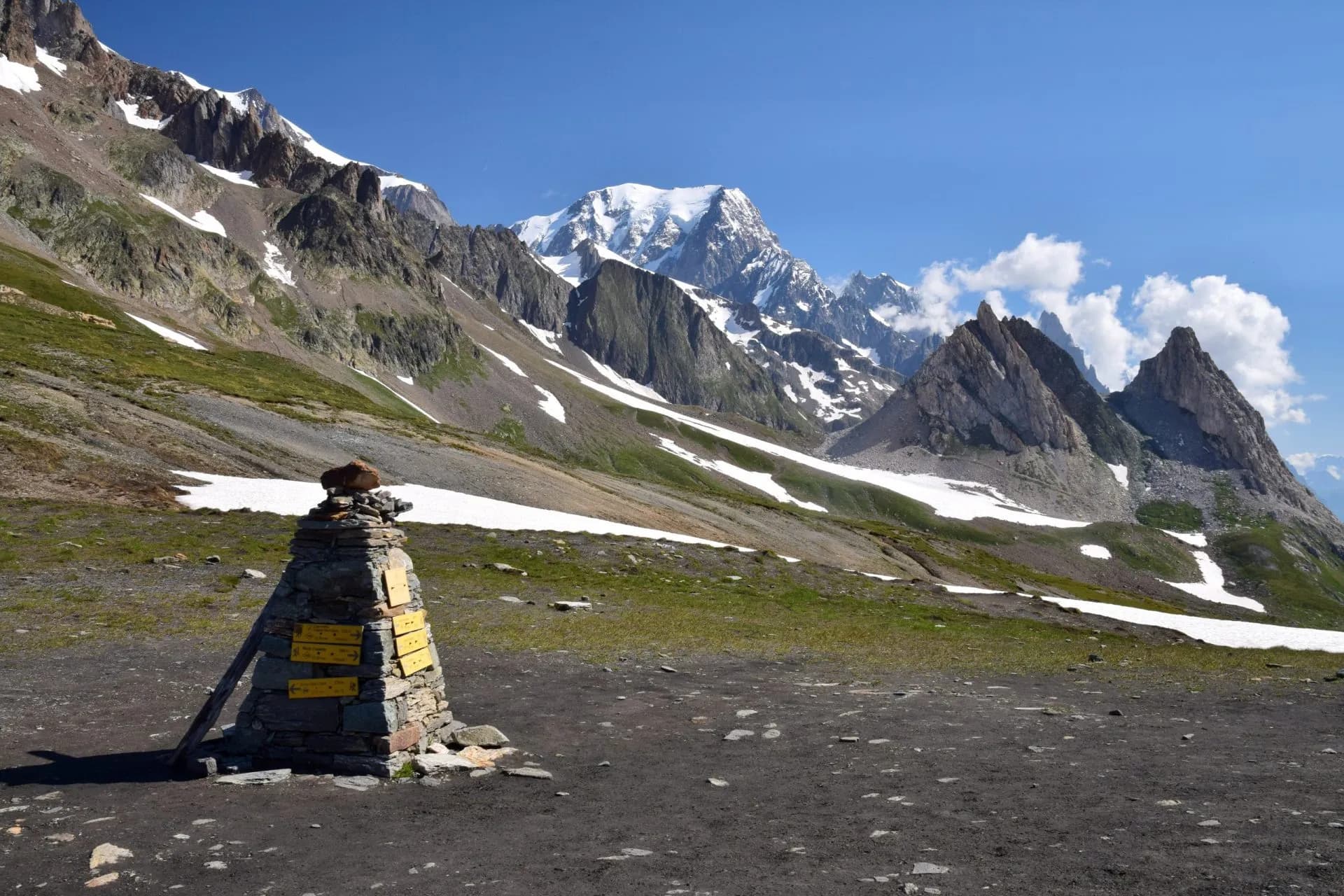 Hiking trail marker cairn with yellow signs against snowy alpine mountains under blue sky at Col de la Seigne.