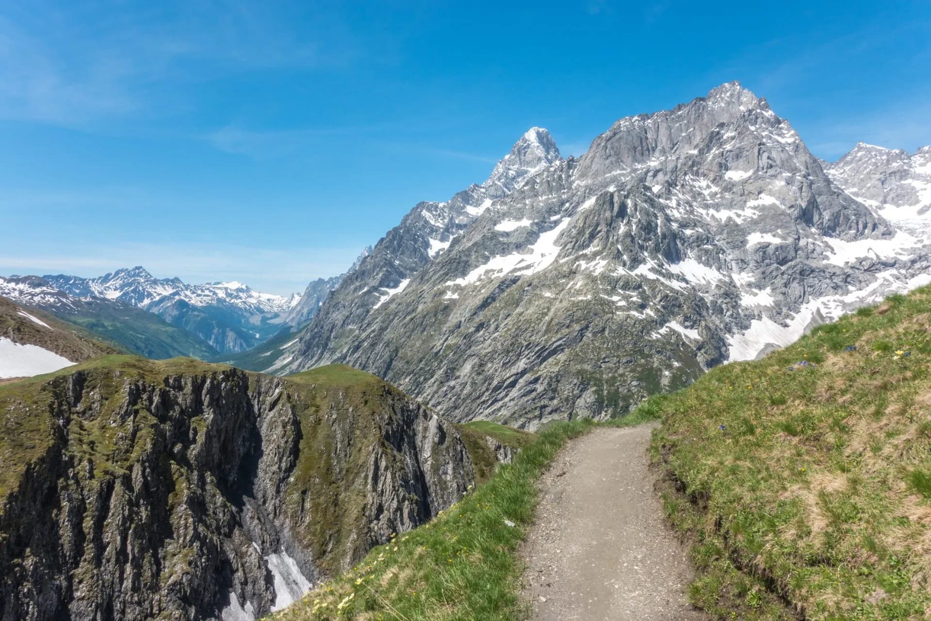 Hiking path along grassy ridge toward snow-capped mountains under blue sky at Grand Col Ferret.