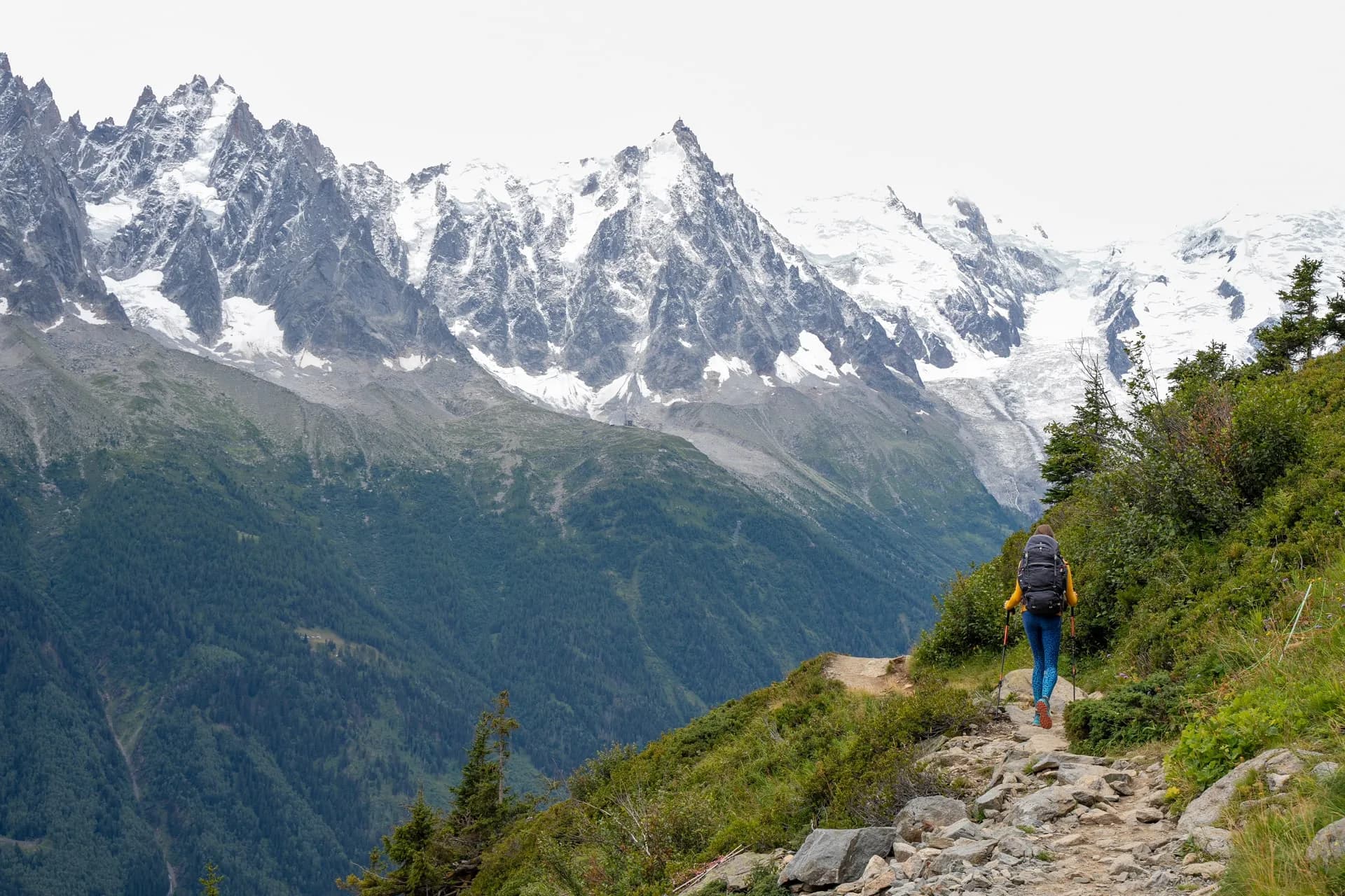 Hiker with backpack walking on rocky trail toward snow-capped mountains and glacier.