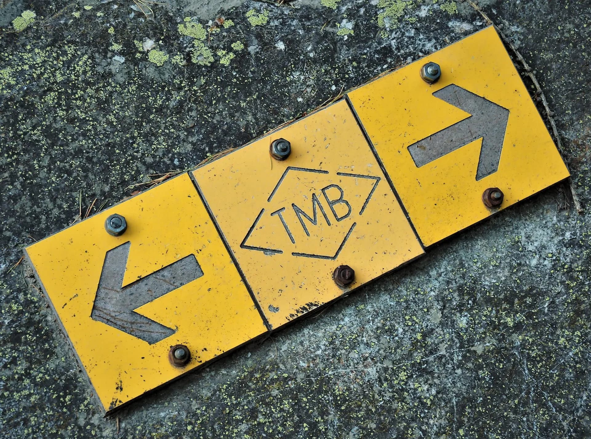 Yellow TMB trail marker sign with left and right arrows bolted onto lichen-covered rock surface.