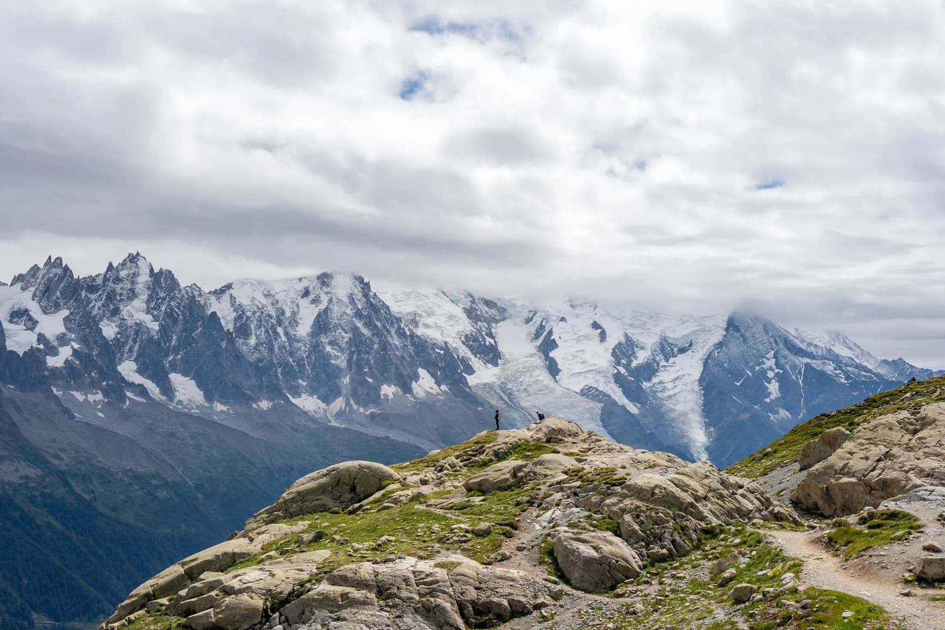Hikers on rocky outcrop with view of massive snow-covered alpine mountains under cloudy sky