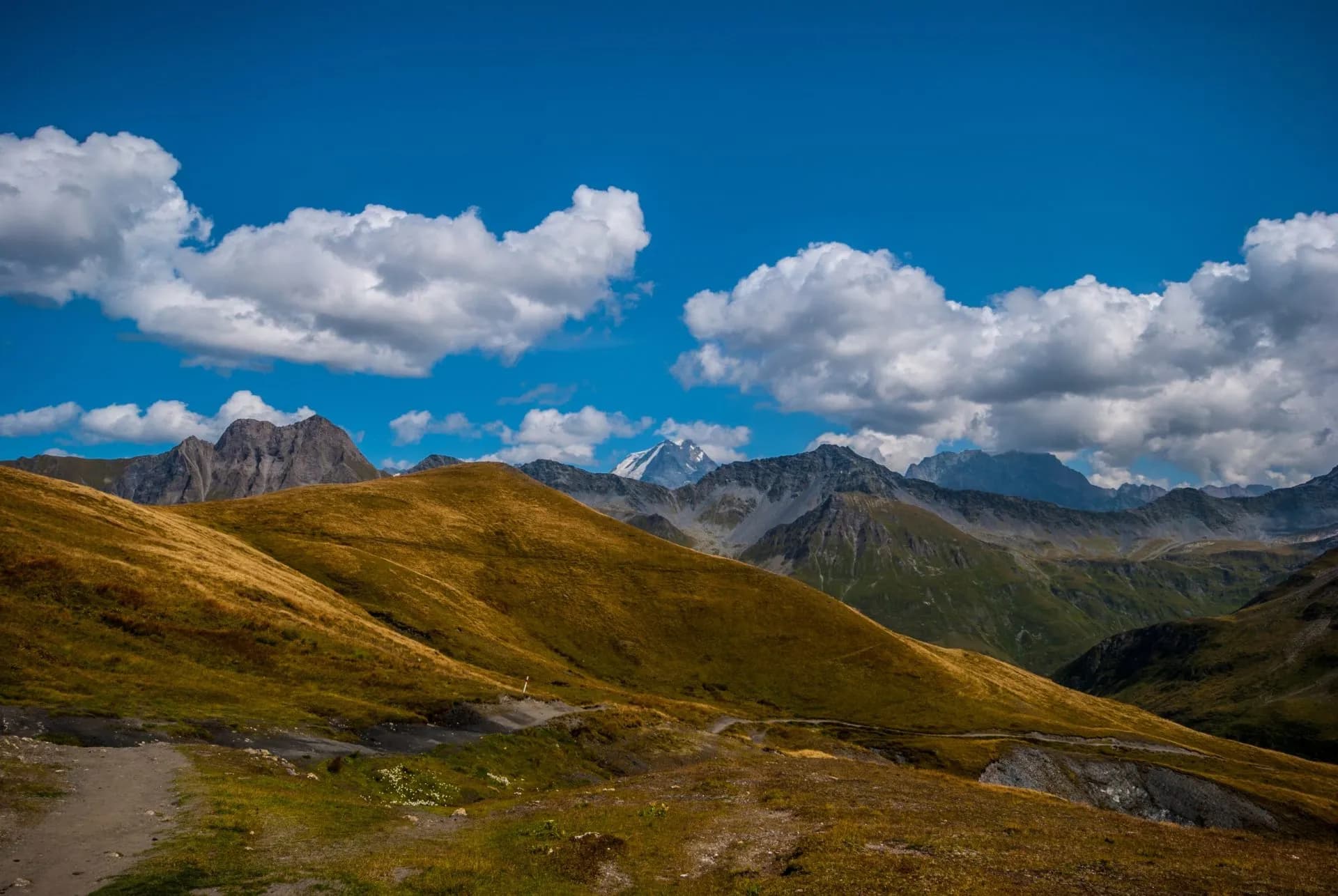 Rolling golden hills and rugged mountains under a bright blue sky with white clouds in La Fouly.