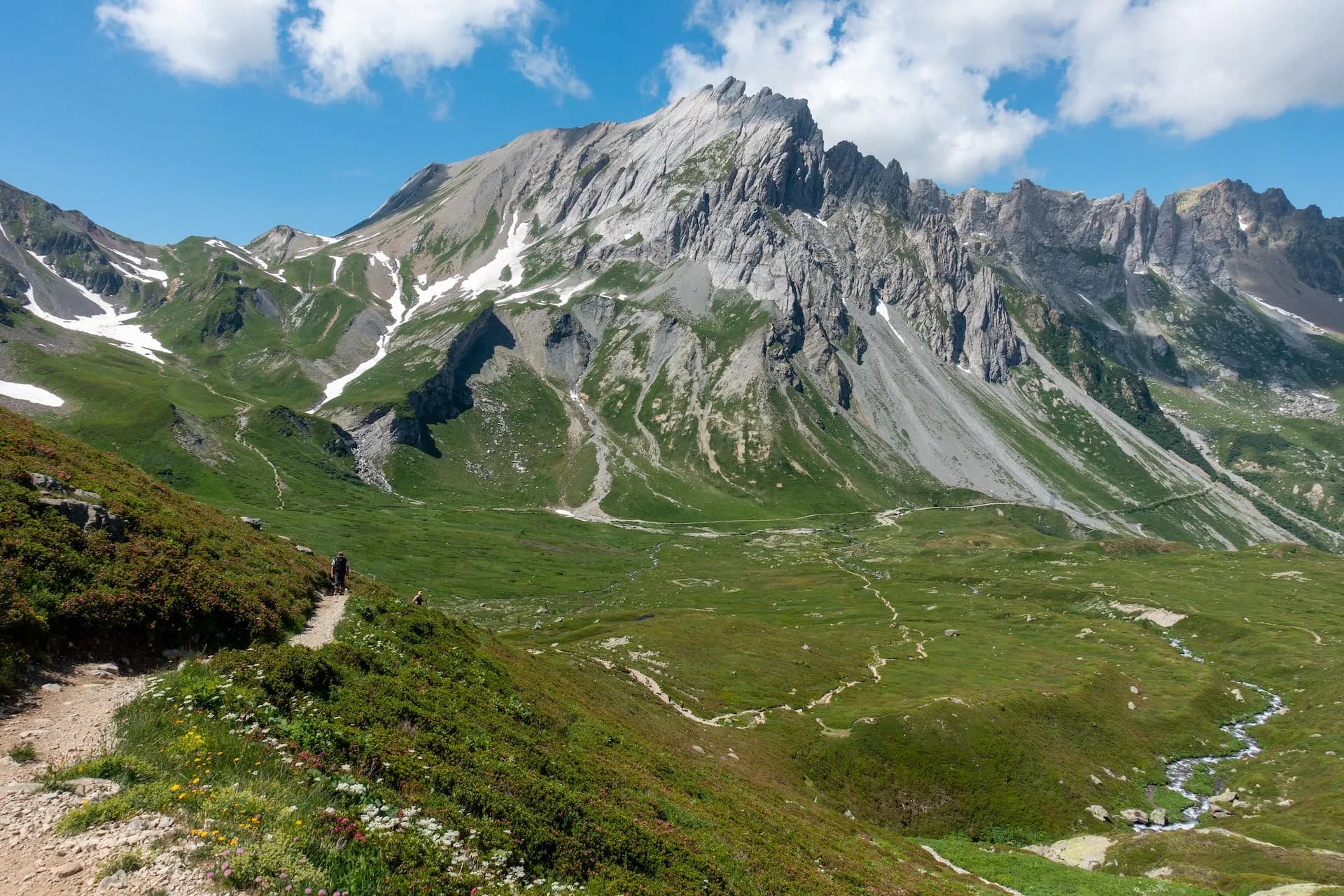 Hikers on trail above green valley with stream, massive rocky mountains, and blue sky at Col du Bonhomme.