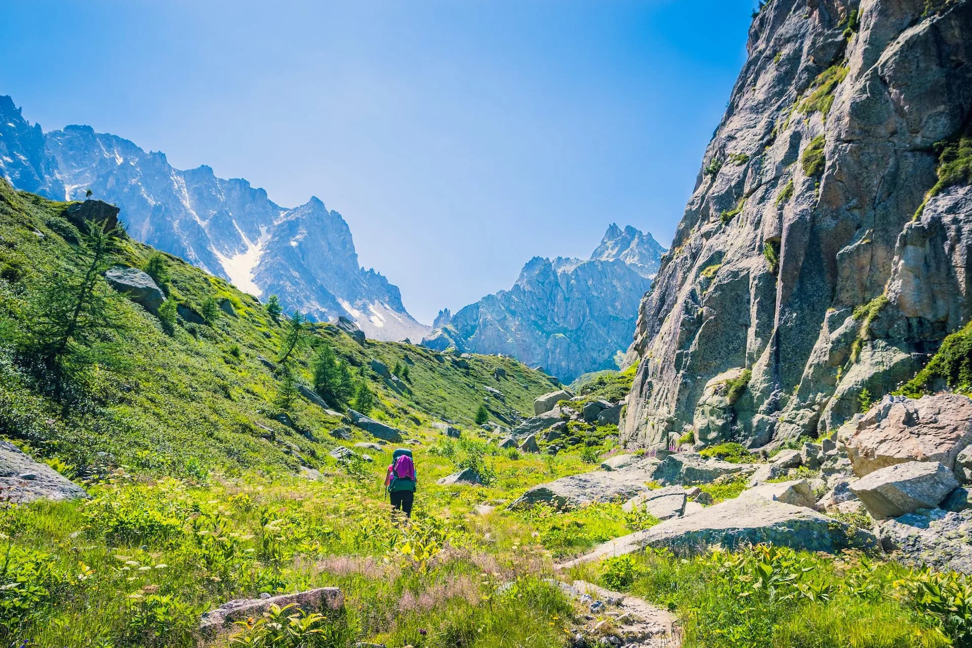 Hiker with backpack walking through green alpine valley toward jagged, snow-dusted mountains.