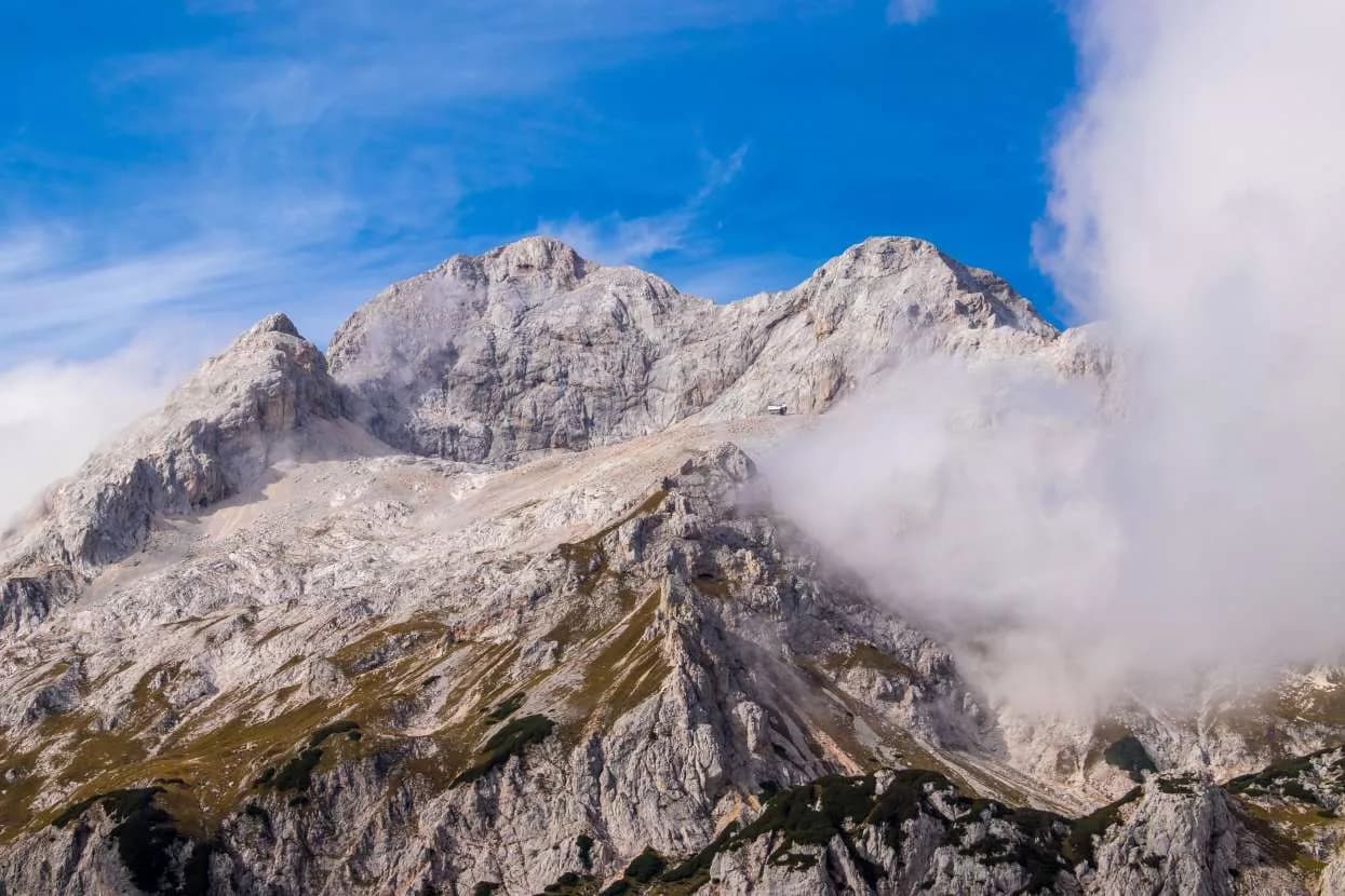 Rocky mountain peaks with sparse vegetation, small hut, and low clouds under a blue sky.