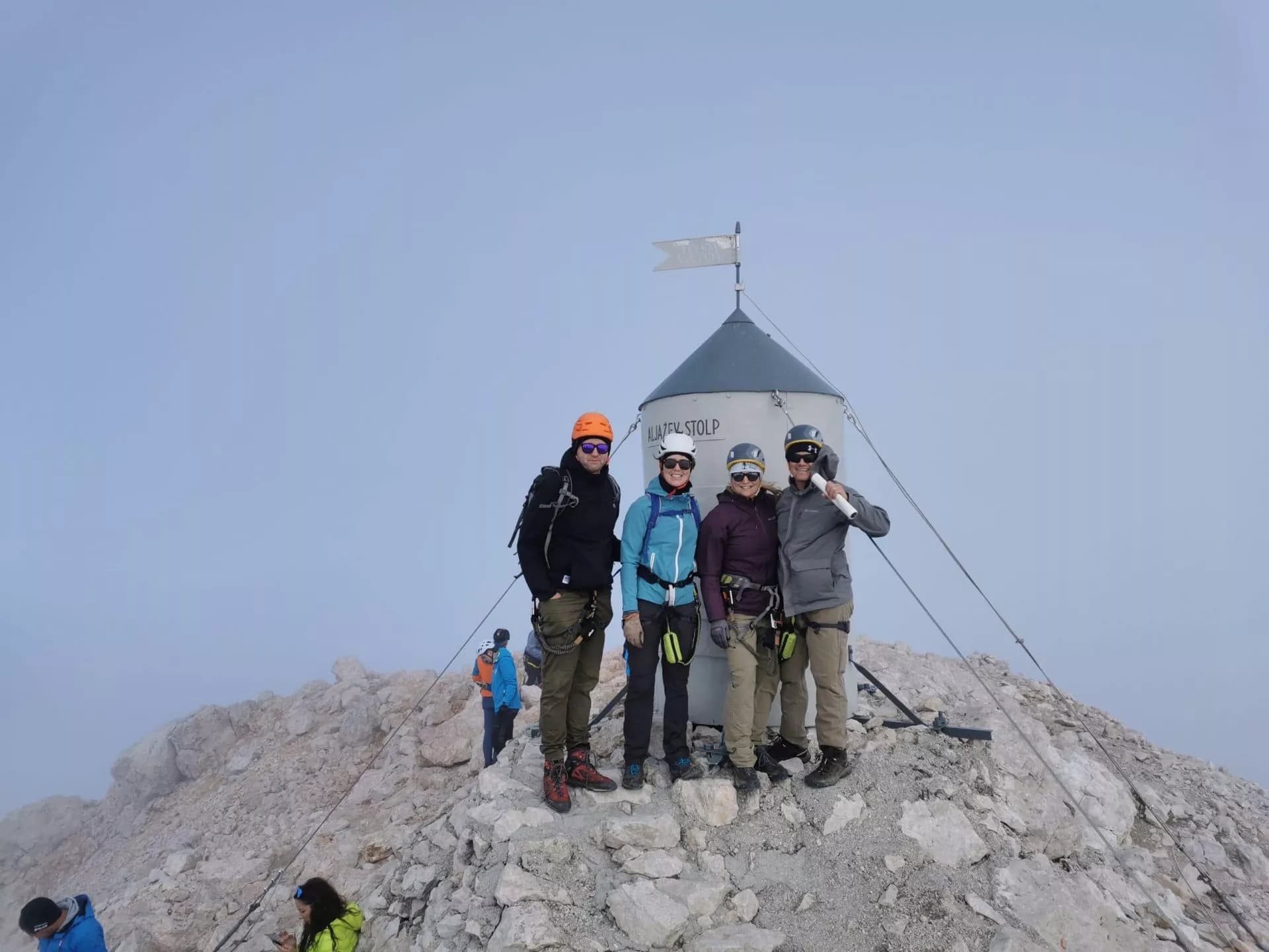Hikers with climbing gear pose by Aljažev Stolp summit marker on rocky mountain peak.