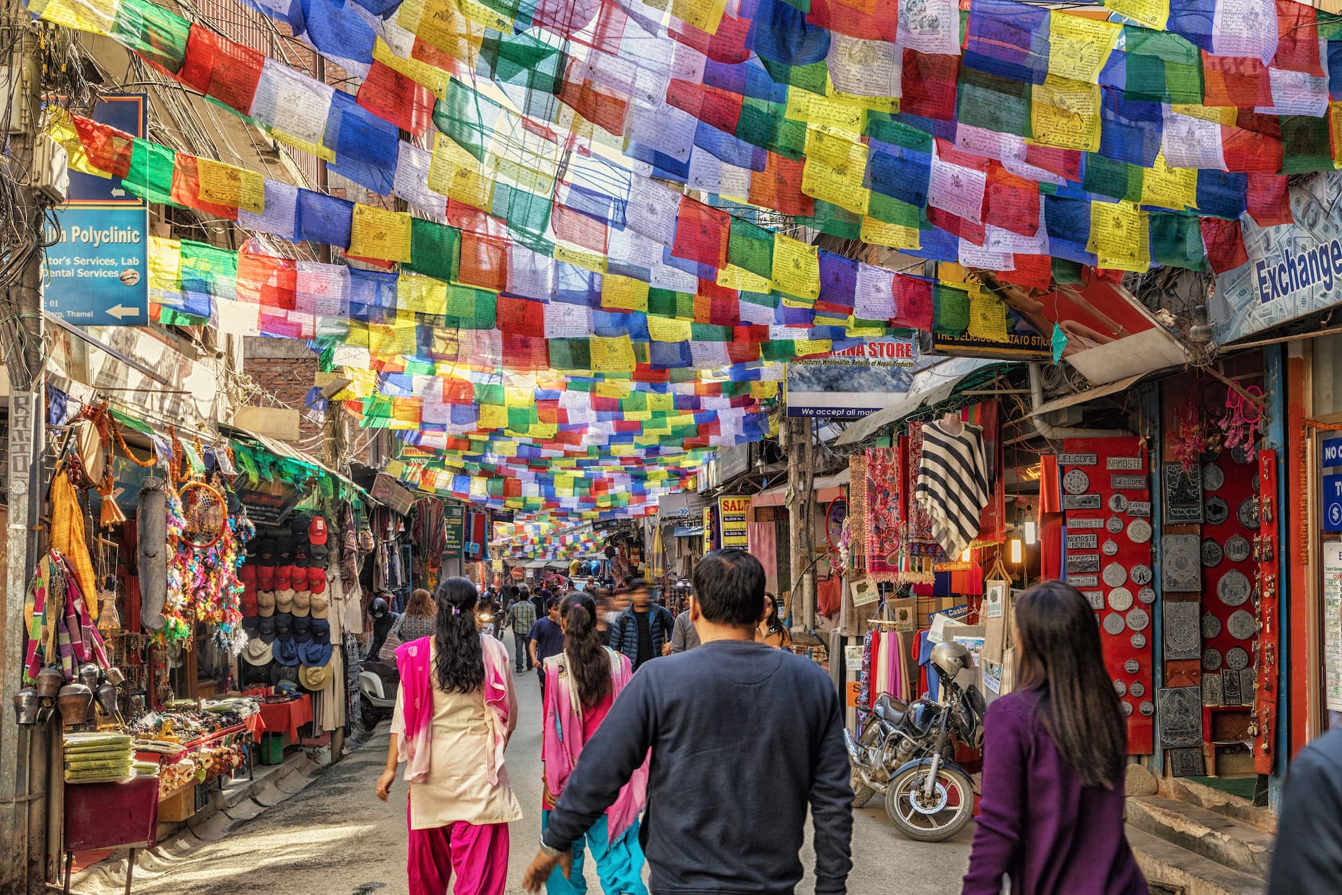 Crowded street market in Kathmandu with colorful prayer flags strung overhead and shops selling goods.