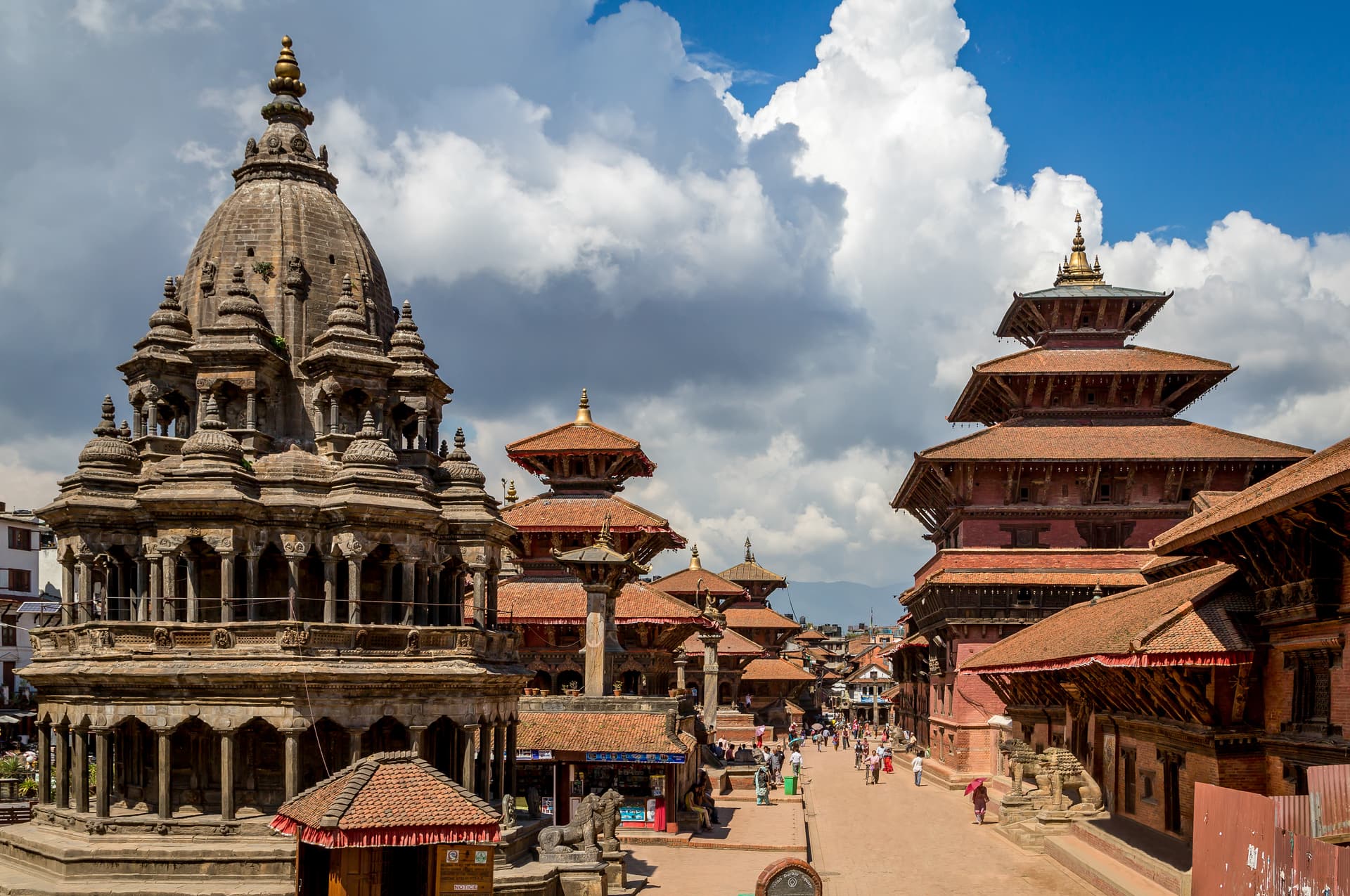Durbar Square with stone temple, tiered brick buildings, and people under a blue sky with white clouds.
