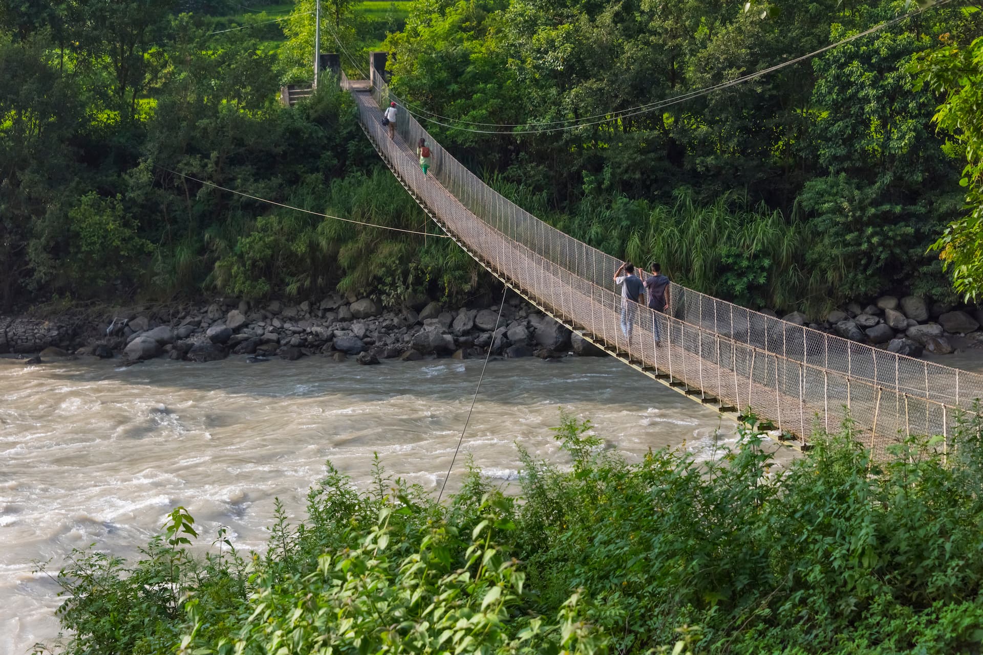 People crossing the Trishuli River suspension bridge over fast-flowing muddy water.