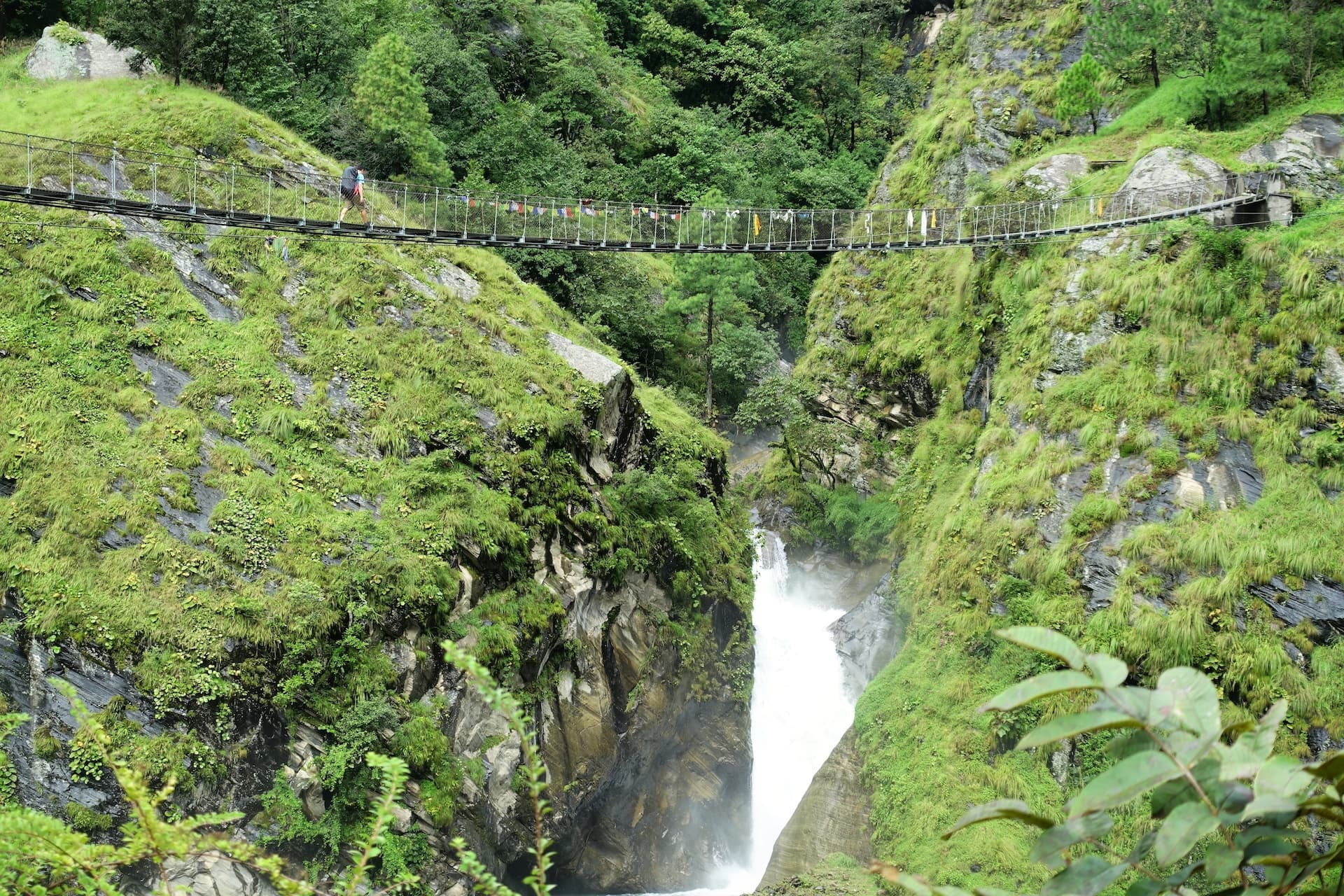 Hiker crosses suspension bridge over deep gorge with waterfall near Jagat.