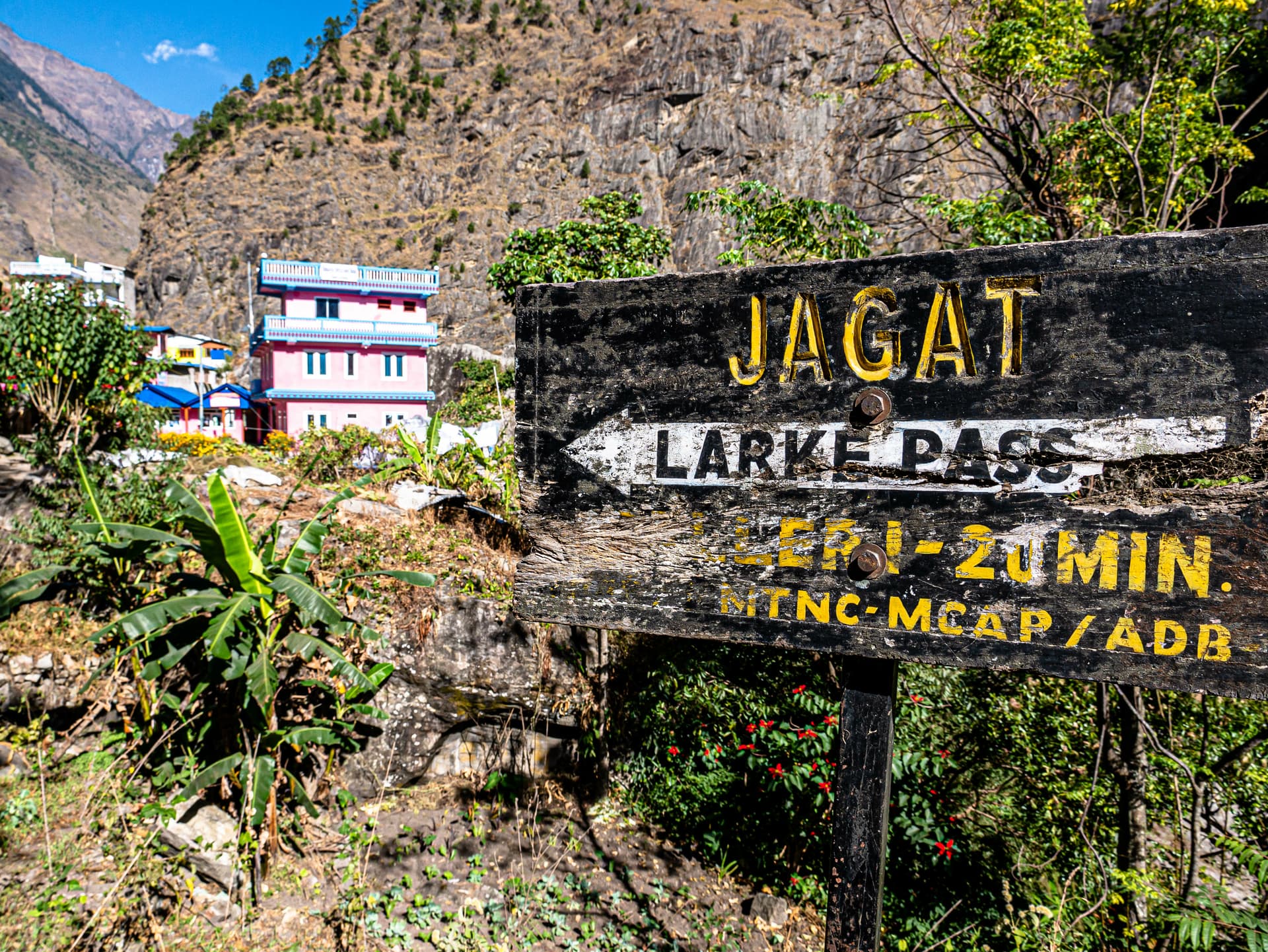 Wooden trail sign for Jagat and Larke Pass in a mountainous setting with colorful buildings.