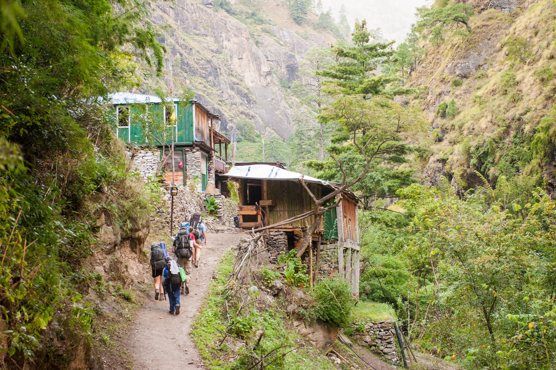 Trekkers hiking on a dirt path past rustic buildings toward Lho village in a steep, green valley.