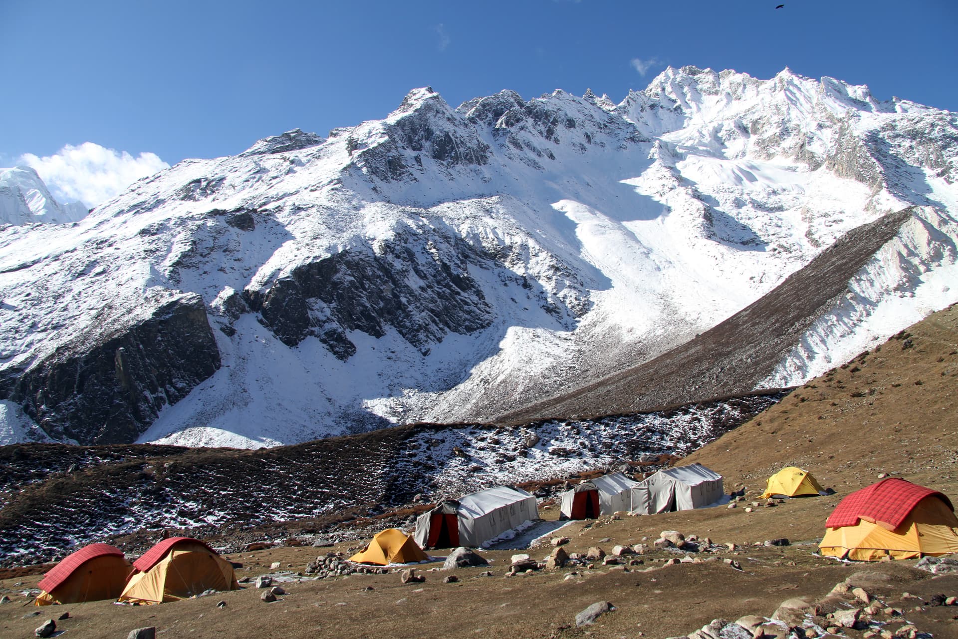 Tents set up at a high-altitude base camp beneath massive snow-covered mountains under a blue sky.
