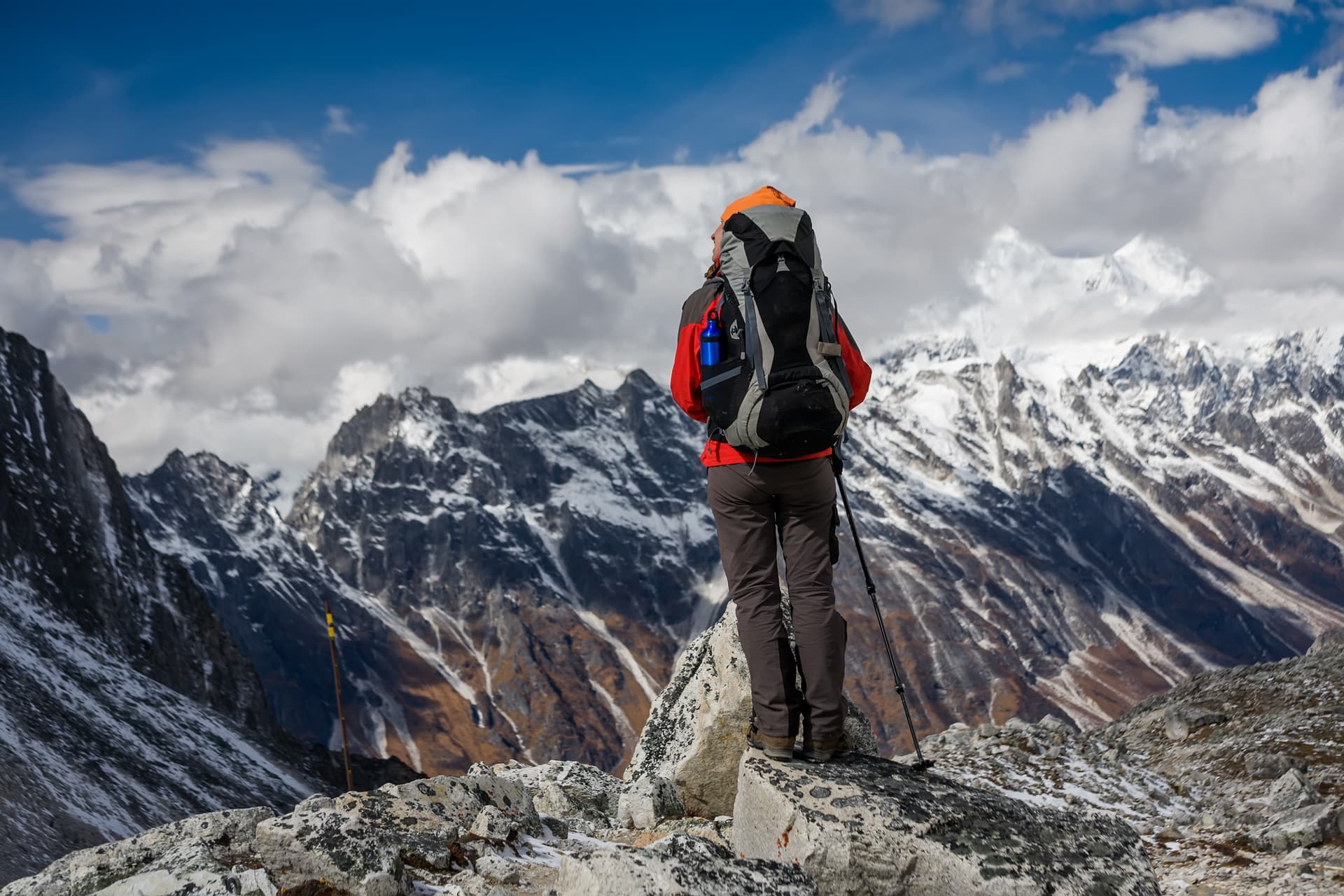 Hiker with backpack standing on rocks overlooking snow-capped mountains under a cloudy blue sky, Larke La Pass Manaslu.
