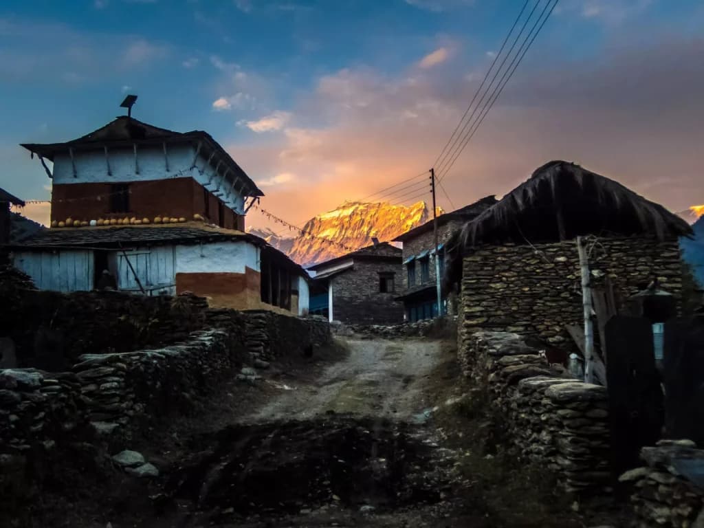 Dharapani village dirt road with traditional stone and wood houses and sunlit mountain peak.